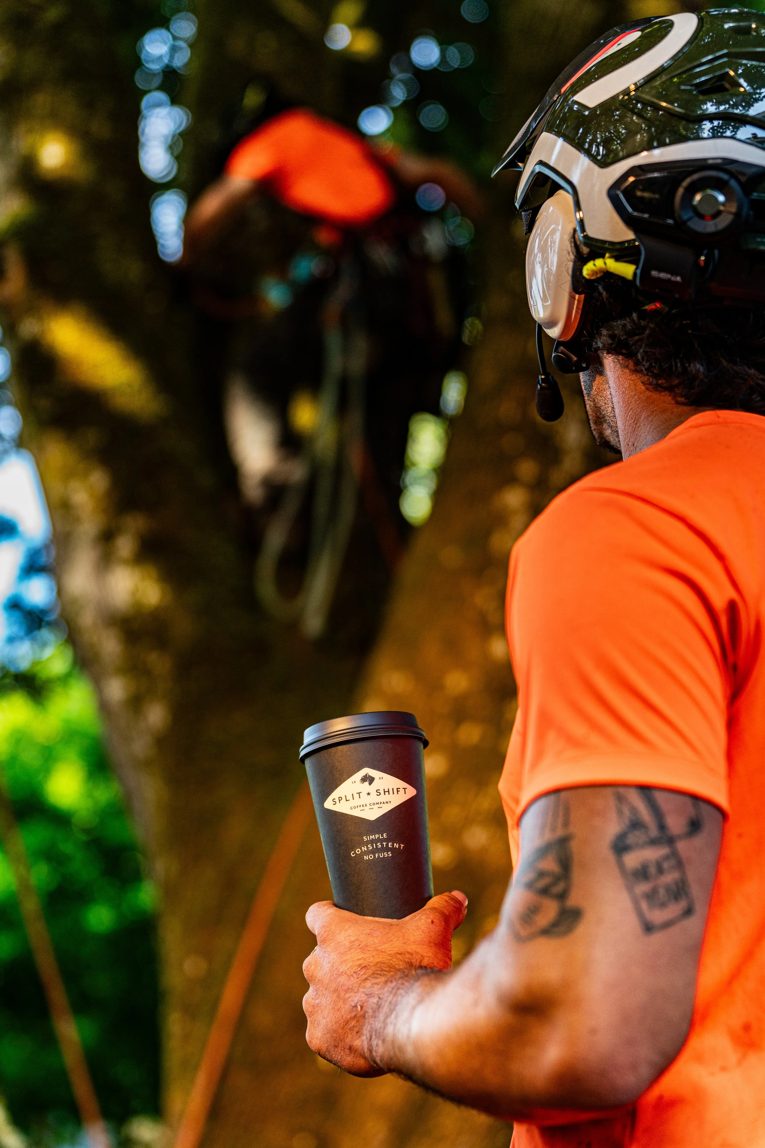 A person wearing an orange shirt and a white helmet is holding a black cup from Split Shift Coffee Company. The person has tattoos on their arm and is outdoors, with trees and blurred lights in the background.