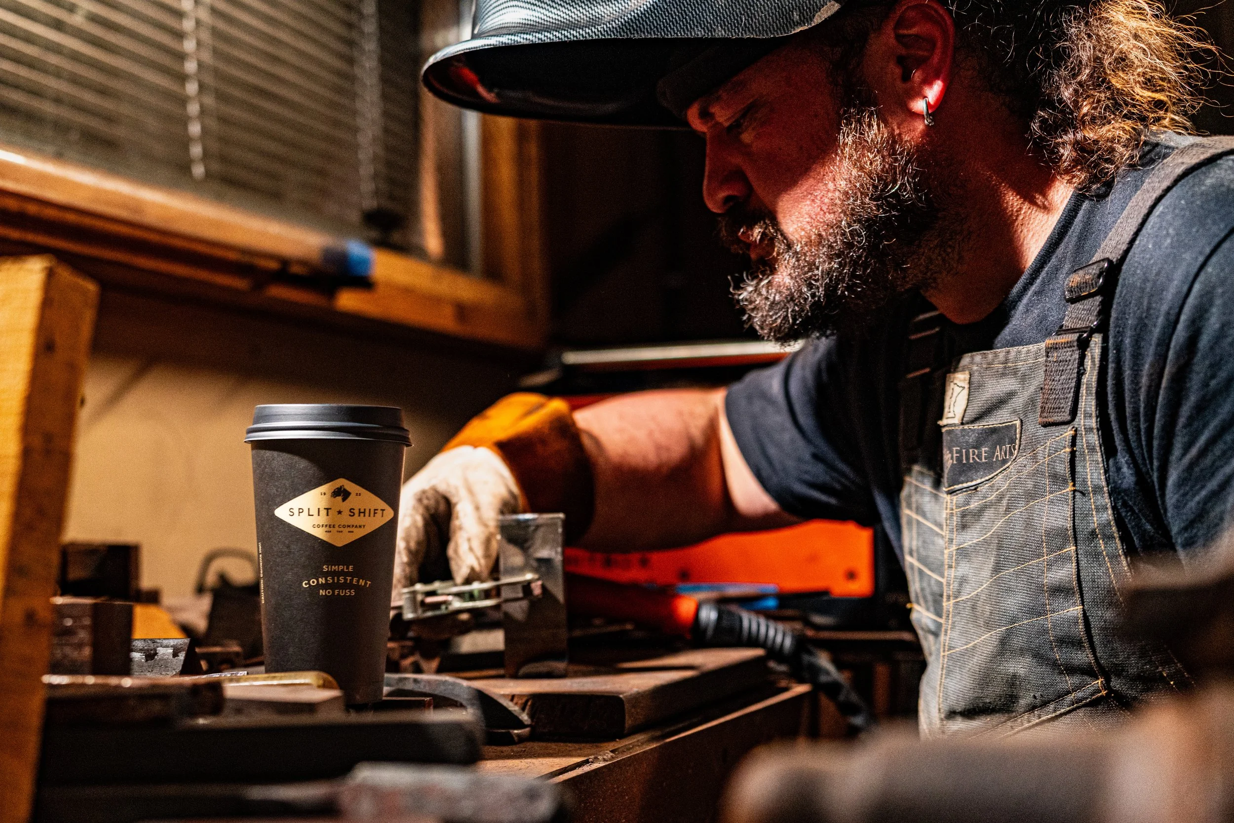 A man wearing a hat, earrings, and a work apron is focused on working on a project at a workbench. There is a to-go coffee cup with a logo on it in the foreground.