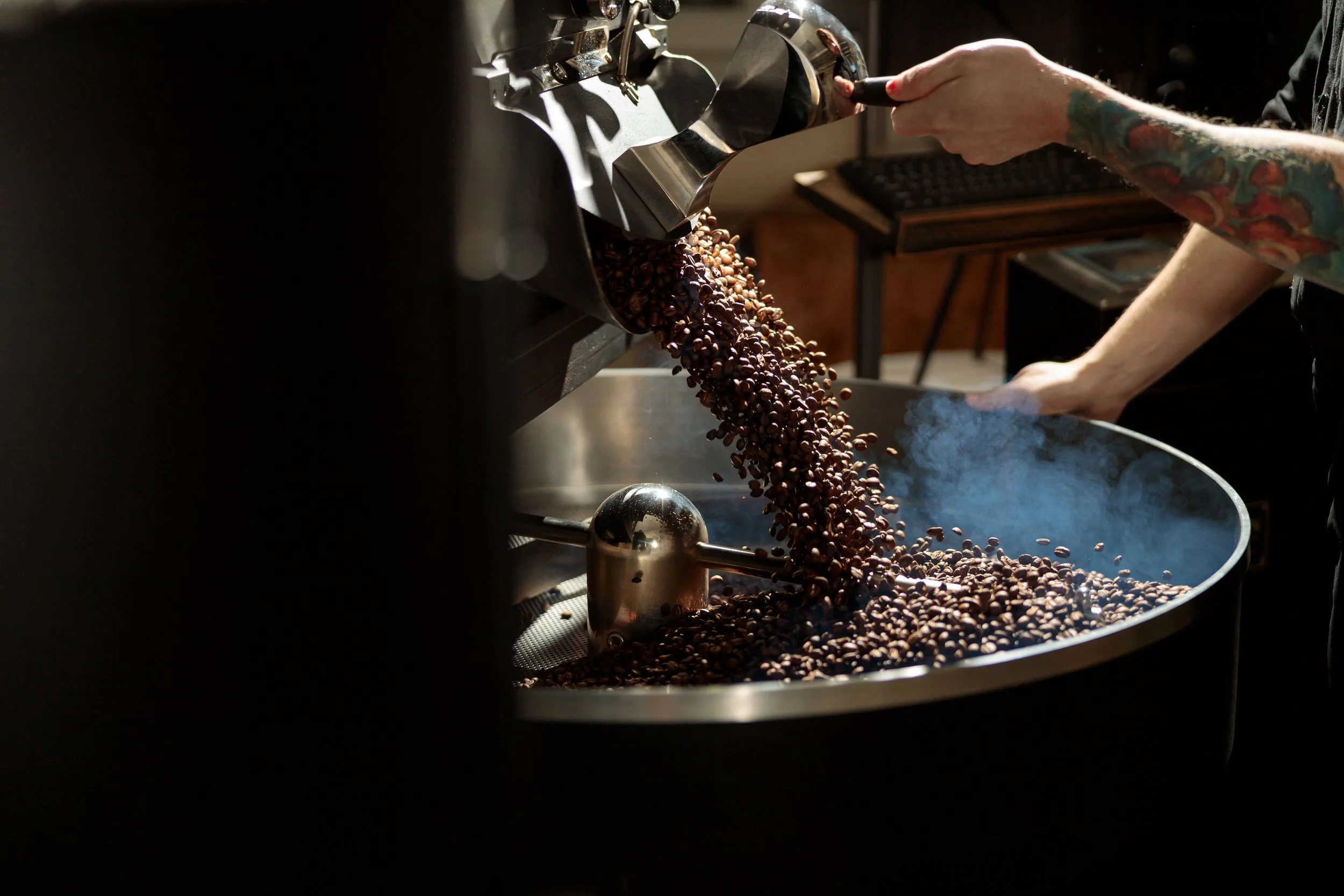 Close-up of a person with colorful tattoos on their arm, using a coffee roasting machine to roast coffee beans.