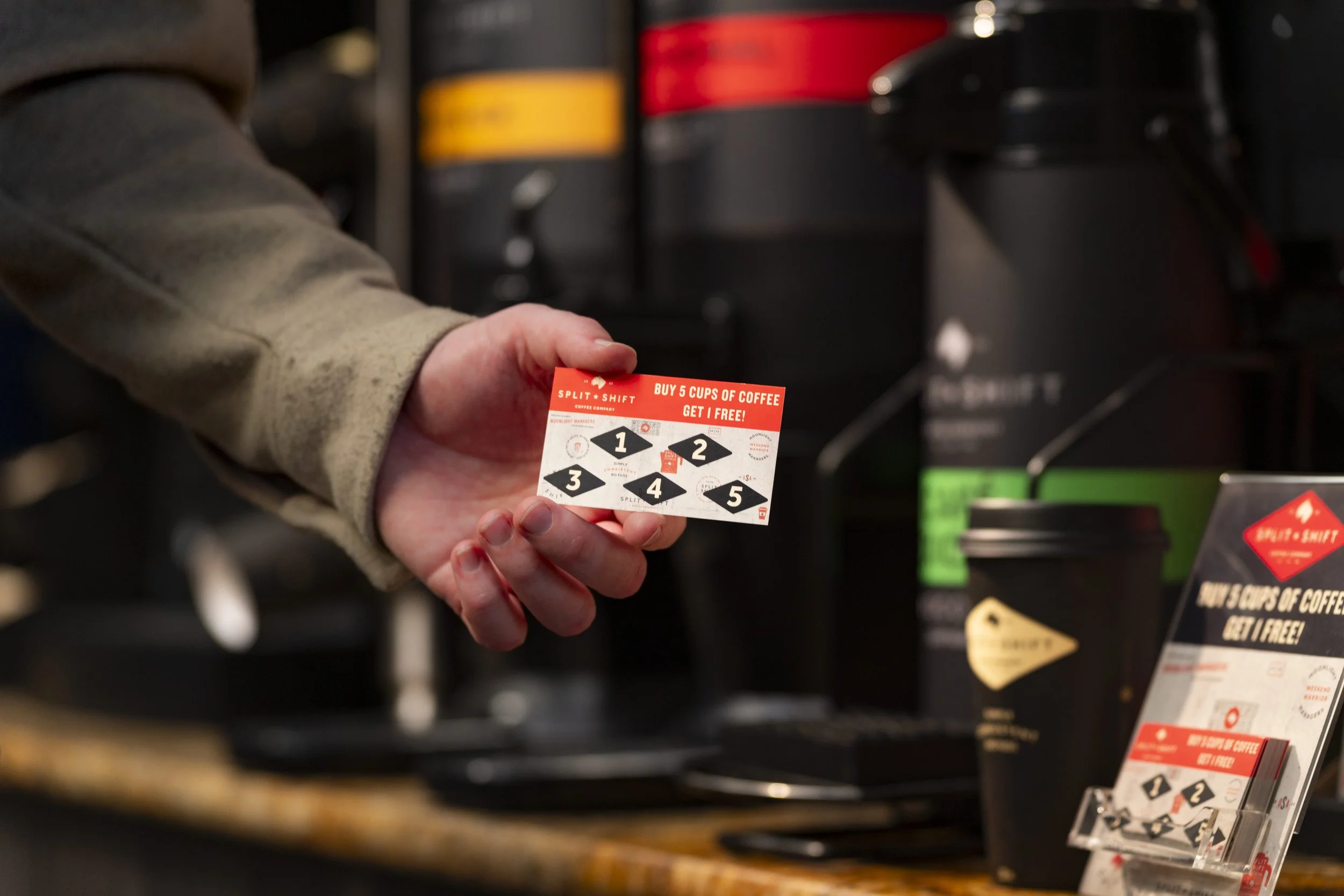 A person holding a promotional card for coffee deals at a coffee shop or convenience store. The card has a red and white design with a dotted pattern and bold text.