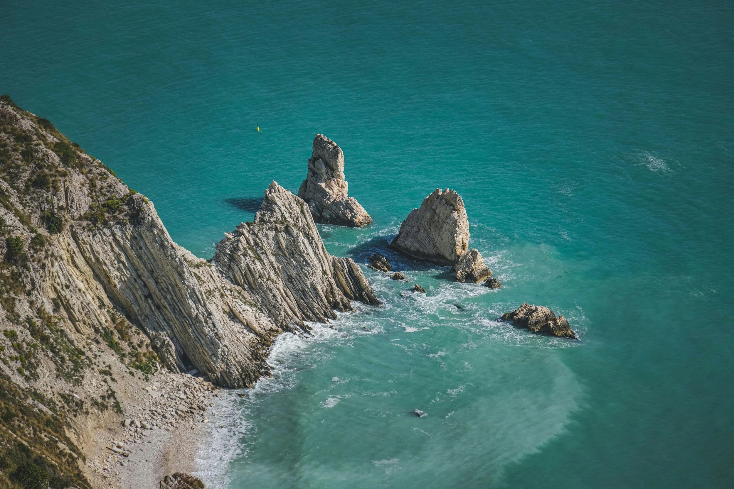 Aerial view of a rugged coastline with tall white rocks protruding from the turquoise sea, with green vegetation on the cliffs.