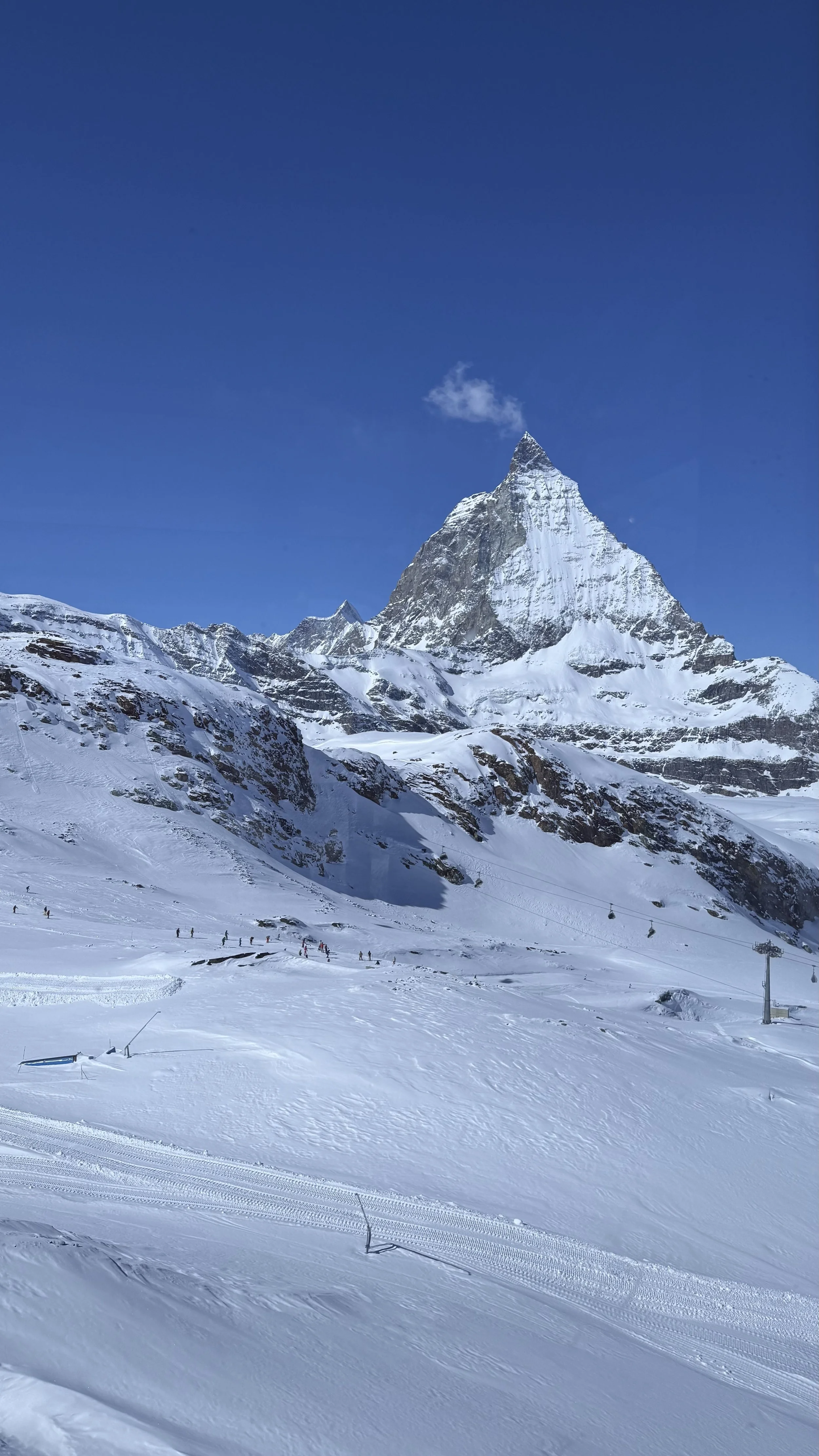 Snow-covered mountain with a sharp peak under a clear blue sky, ski slope with skiers and ski lifts in the foreground.