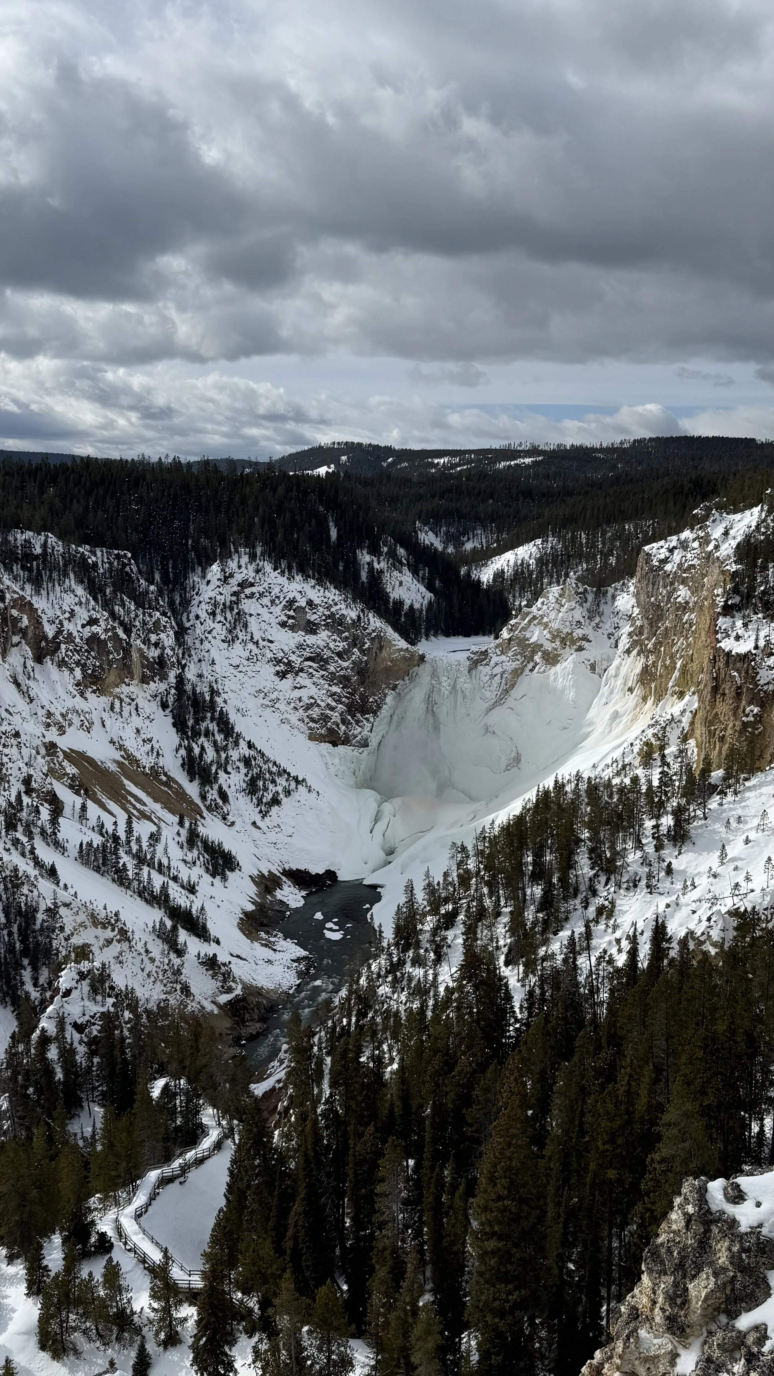 Snow-covered mountains and a waterfall with ice and snow in a forested valley under cloudy sky.