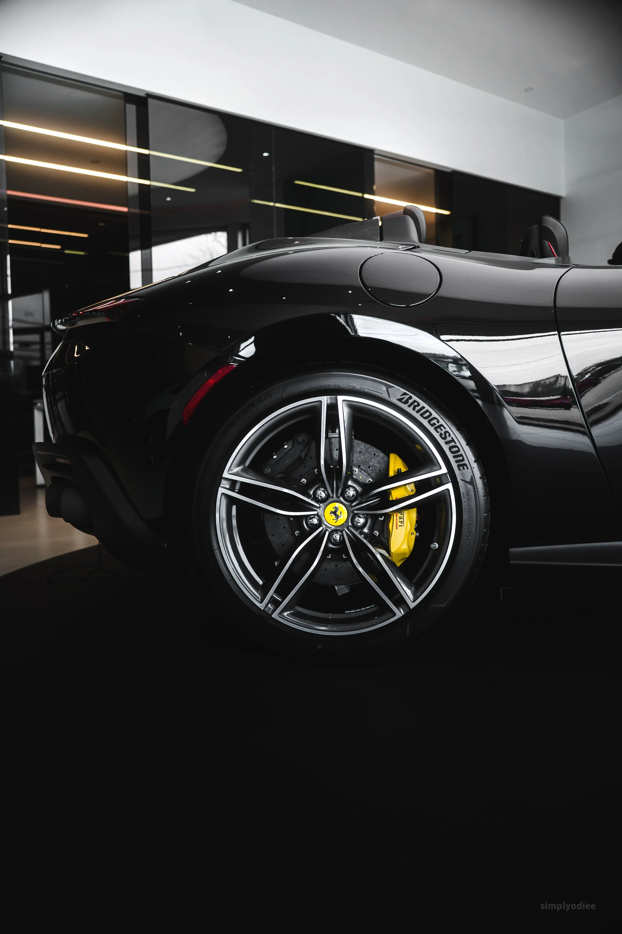 Close-up of the rear wheel and side profile of a black Ferrari convertible sports car, featuring yellow brake calipers and Bridgestone tires, inside a modern showroom with glass walls and bright ceiling lights.