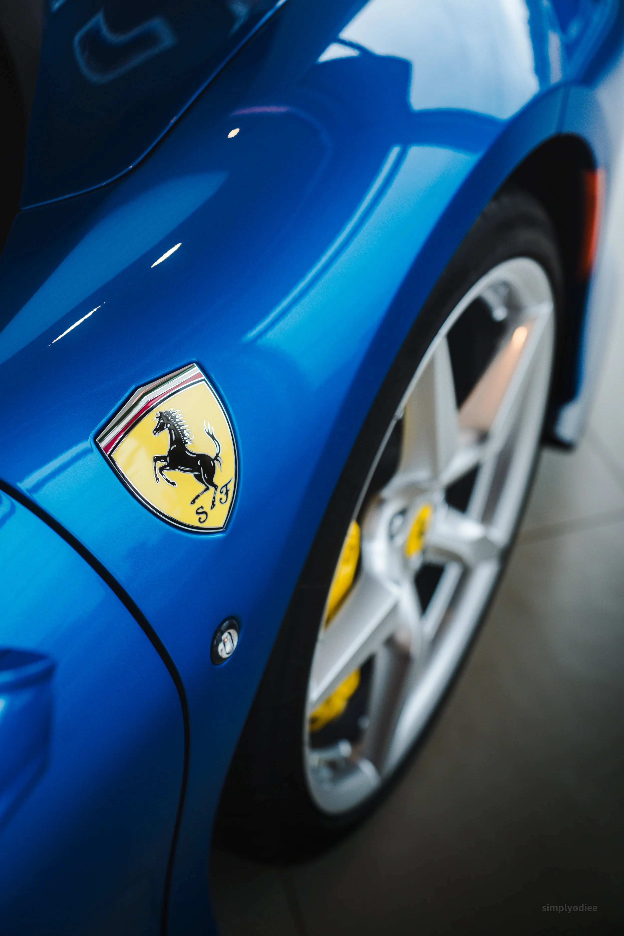 Close-up of a blue Ferrari sports car with a Ferrari emblem on the front. The wheel and part of the car's body are visible.