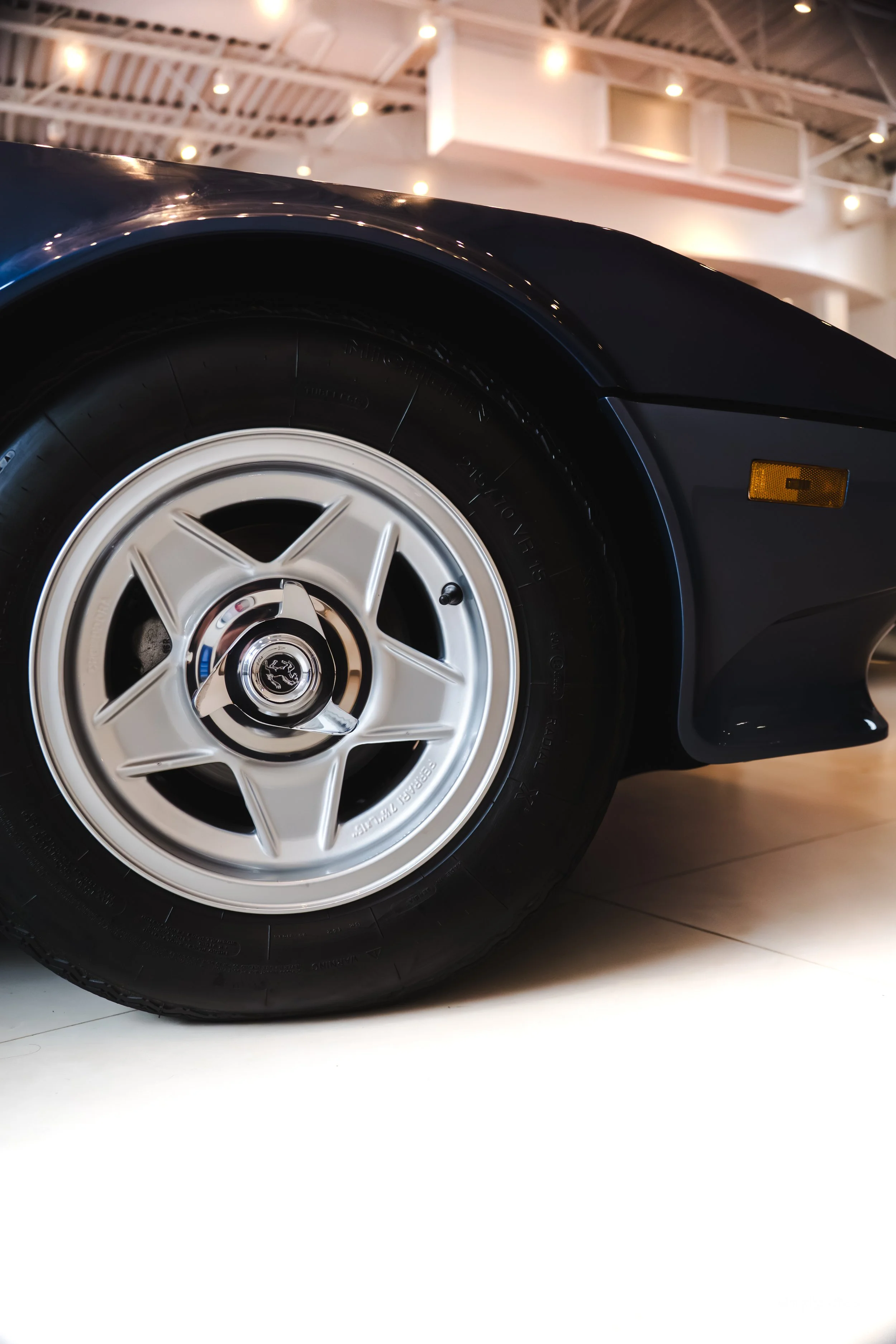 Close-up of a classic black sports car wheel with silver alloy rim on a polished floor in an indoor showroom.