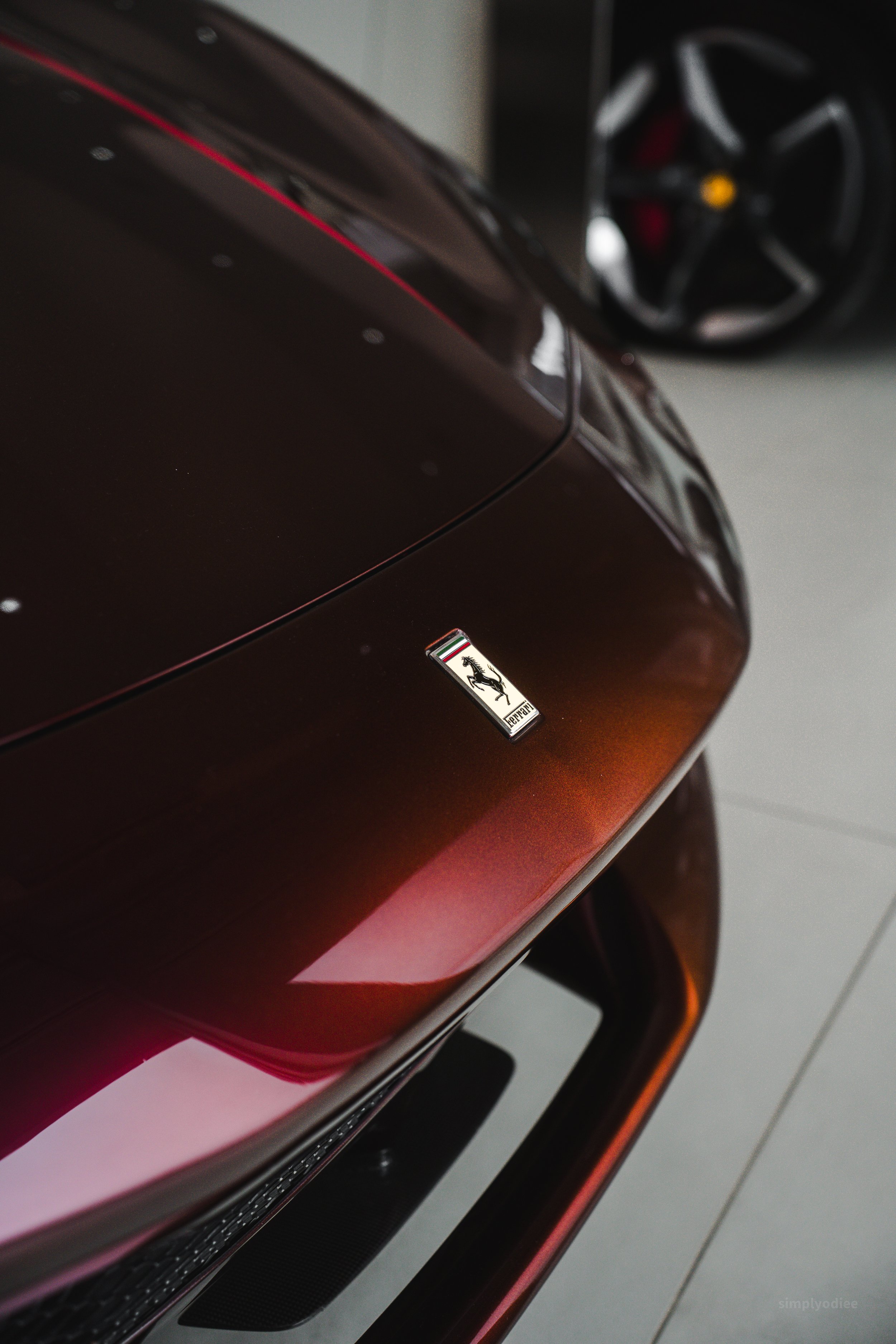 Close-up of a Ferrari logo on the front of a maroon sports car in a garage, with a black car wheel in the background.