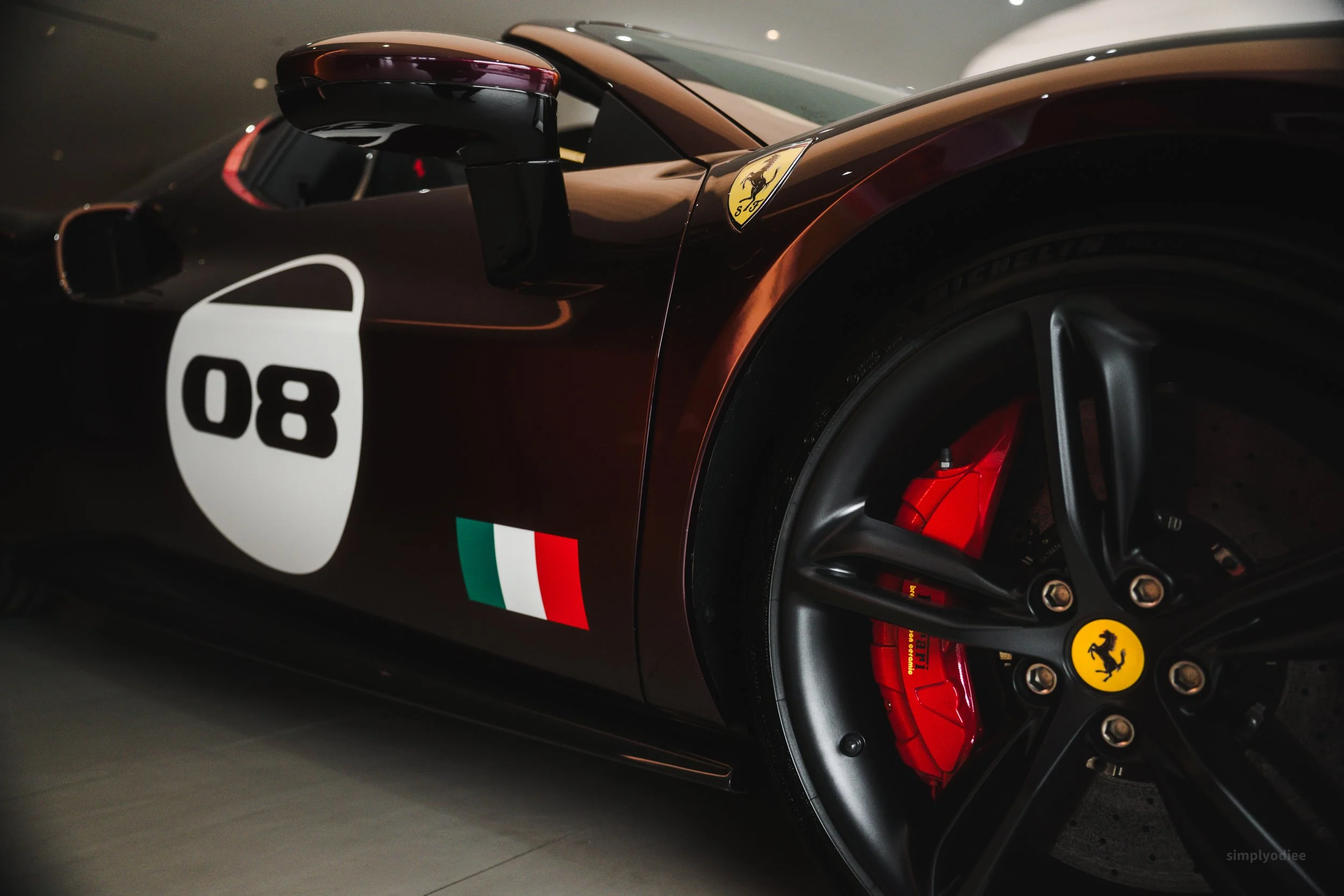 Close-up of a dark bronze Ferrari race car with the number 80 on the side, a Ferrari emblem on the front fender, red brake calipers, and an Italian flag decal.