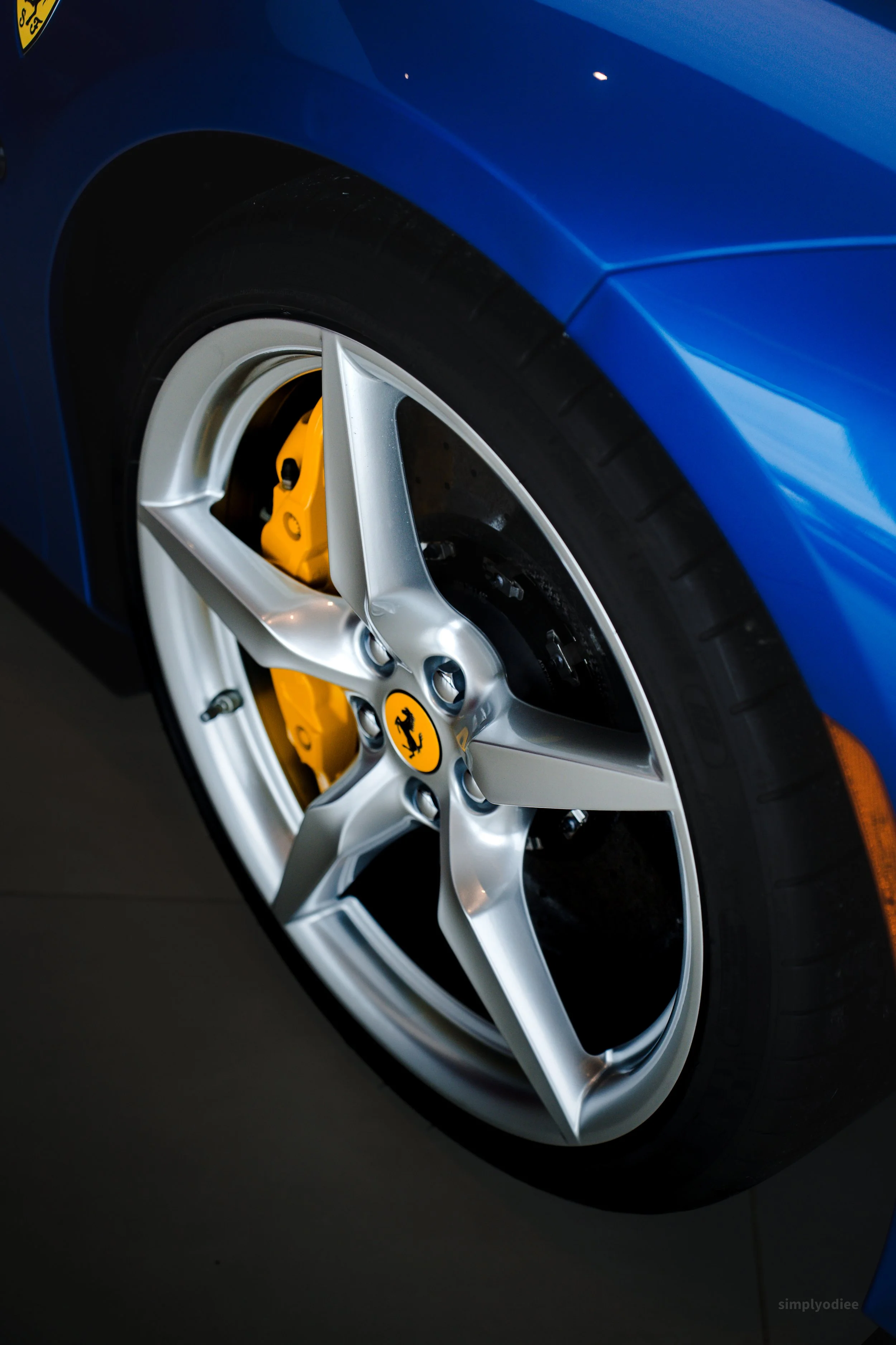 Close-up of a blue Ferrari sports car wheel with a silver alloy rim, yellow brake caliper, and black tire.