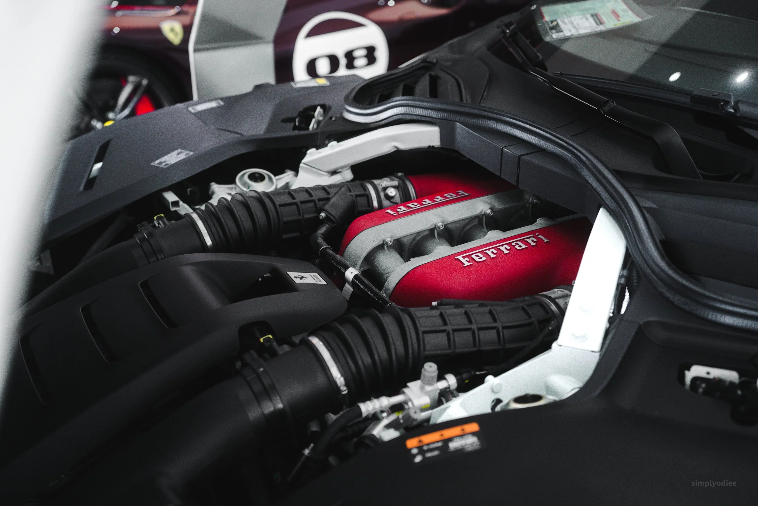 Close-up of a Ferrari car engine with a red intake manifold labeled 'Ferrari' and 'V8', surrounded by black hoses and engine components.