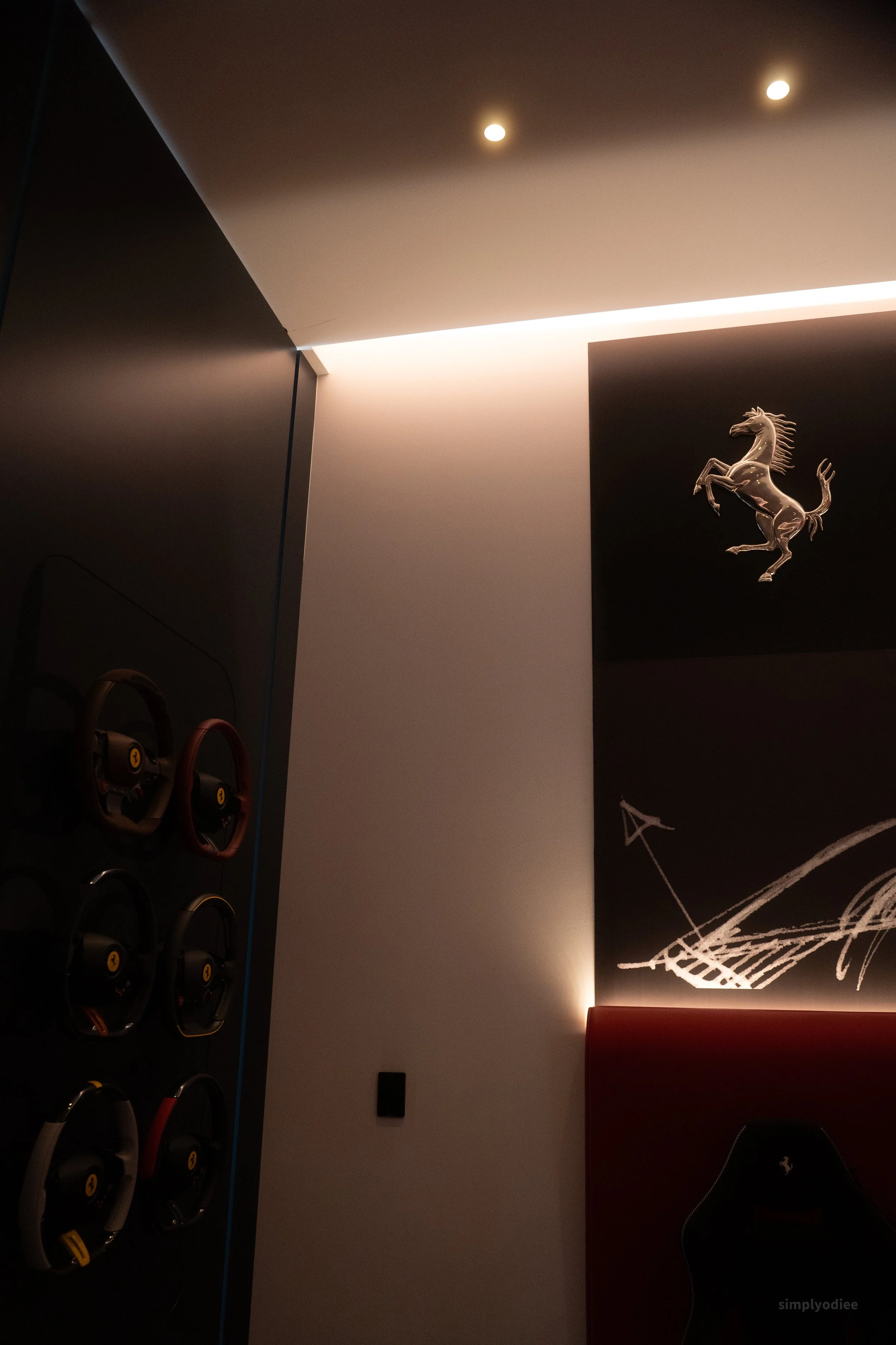 Interior room with a wall-mounted display of six Ferrari car steering wheels on the left, a black wall with a mounted silver horse Ferrari emblem, and a wall with a sketch of a racing car and an arrow pointing to it. The ceiling has two small recesse