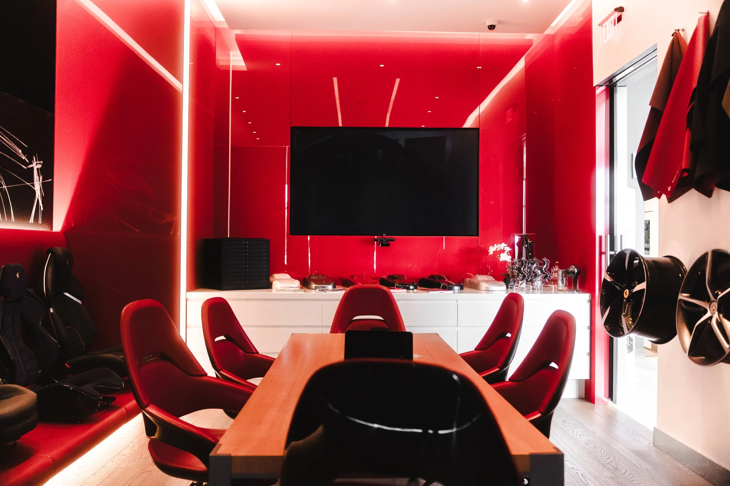 Conference room with red walls, a large television, a wooden table surrounded by six red chairs, and a white cabinet with electronics and decorative objects.