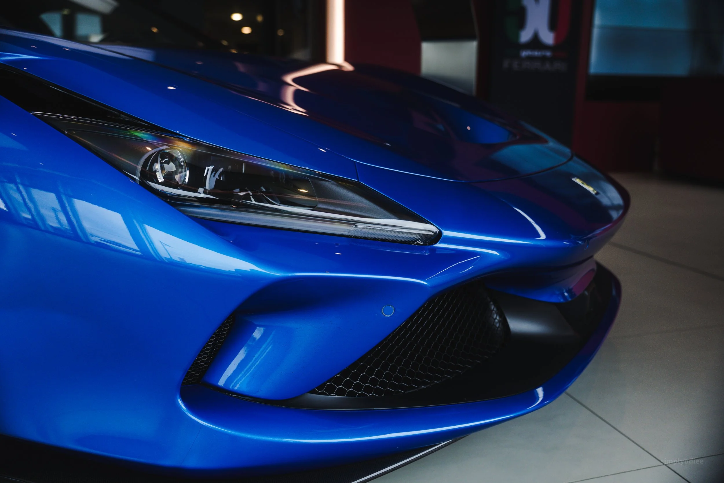 Close-up of a blue sports car's front end, showing the headlight, hood, and black lower grille.