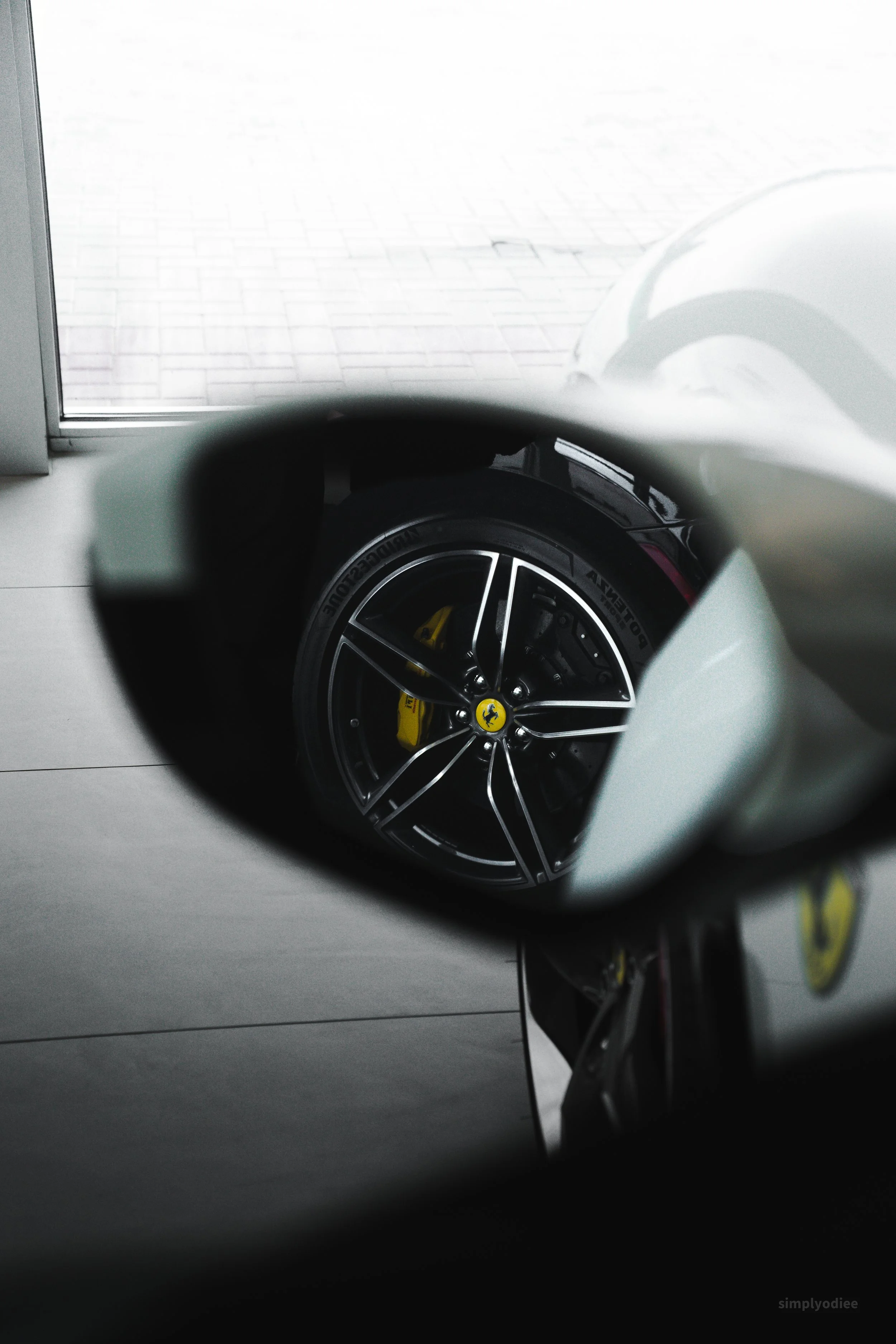 A white sports car, showing a rear view through a side mirror, highlighting the wheel with a red brake caliper and yellow Ferrari logo.
