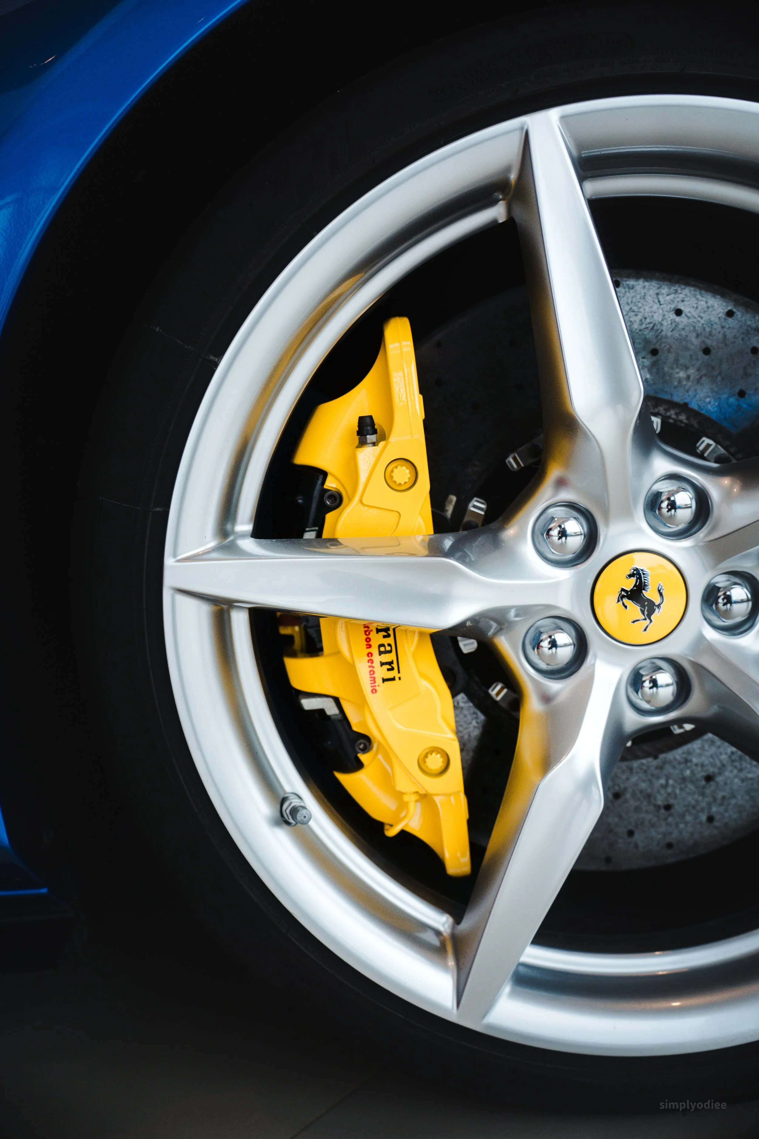 Close-up of a Ferrari car wheel featuring a silver alloy rim, yellow brake caliper, and Ferrari logo on the center cap.