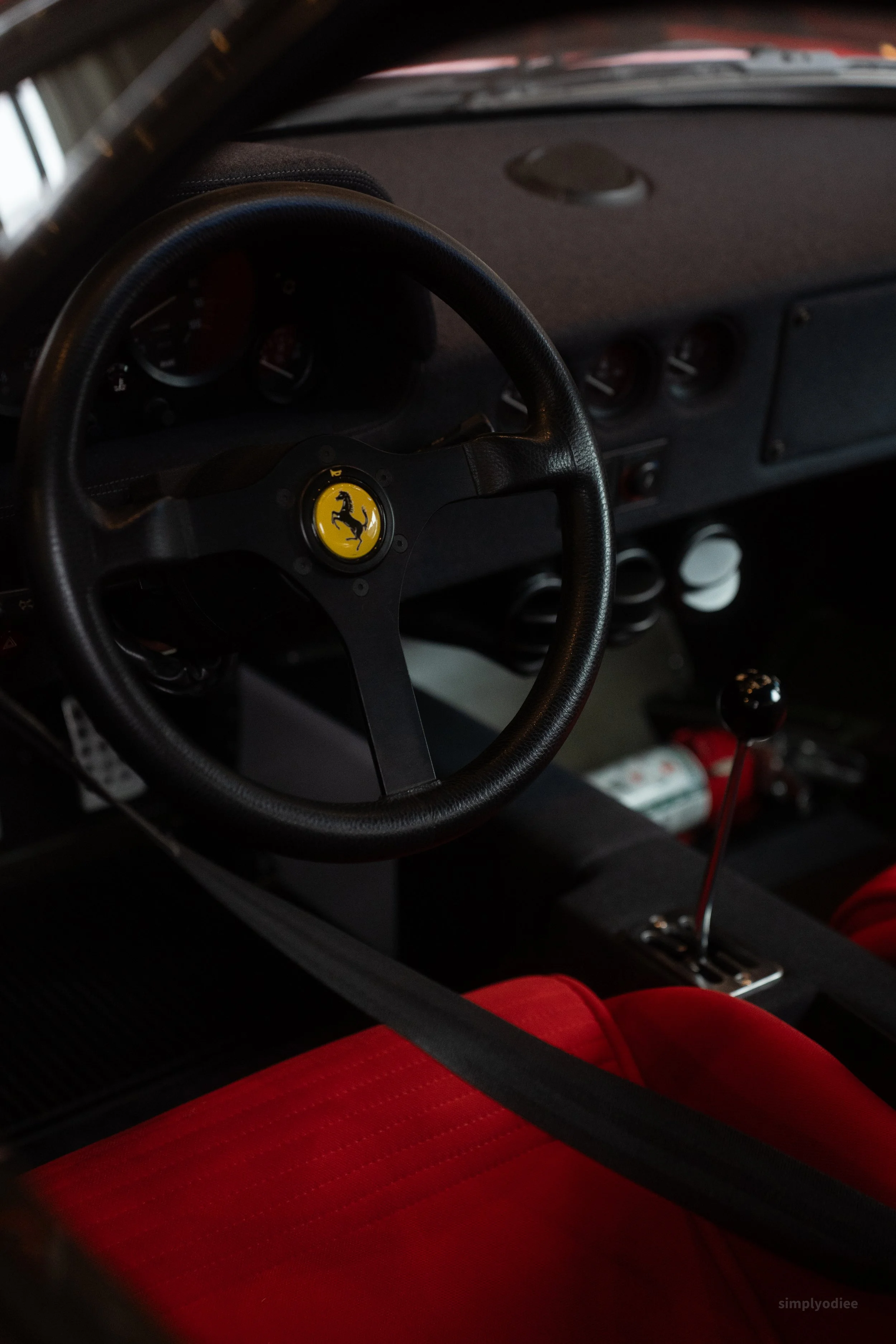 Inside the cockpit of a Ferrari race car, showing a black steering wheel with the yellow Ferrari logo, red racing seat, gear shift, and dashboard instruments.