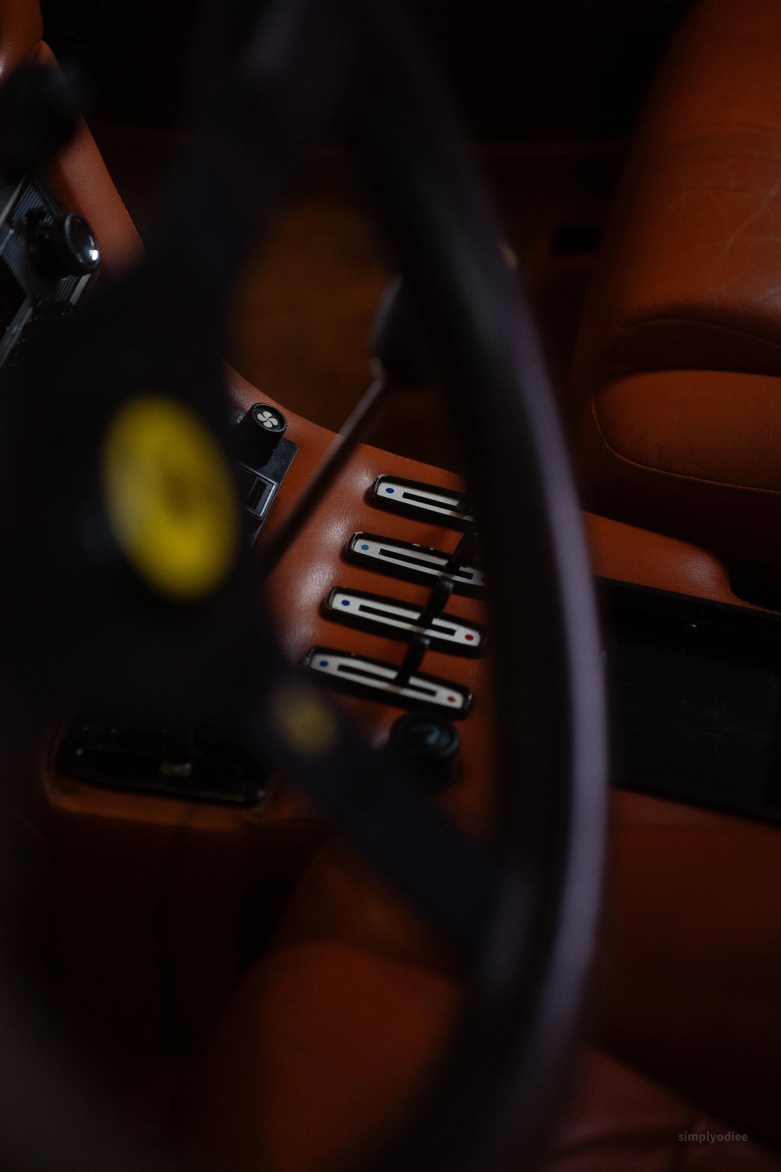 Close-up of a vintage Ferrari steering wheel, dashboard, and gear shift lever, with a leather seat in the background.