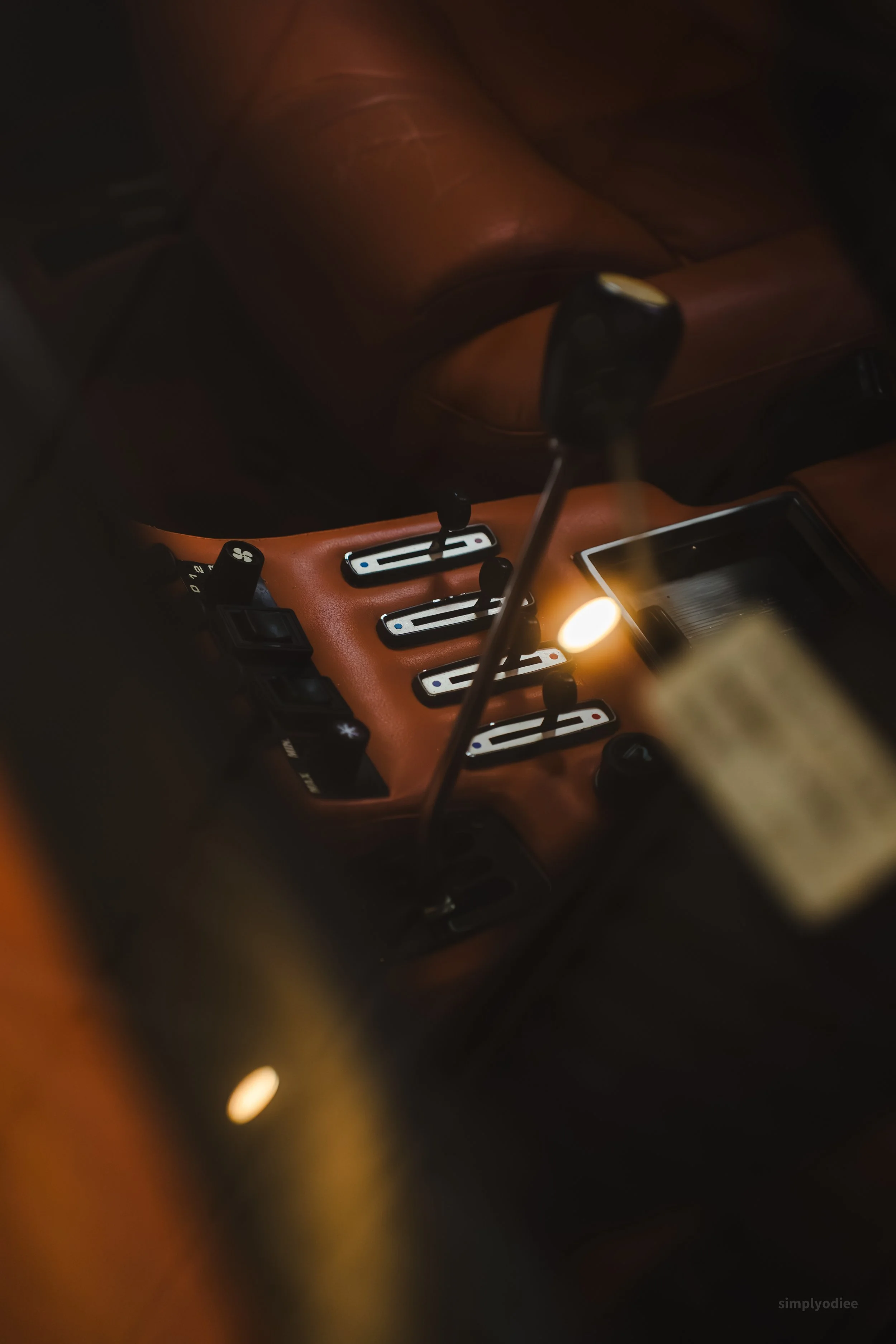 Close-up of the interior of a vintage aircraft cockpit, showing control levers with black knobs, a small dashboard light, and a partially visible screen or instrument panel, surrounded by brown leather seats.