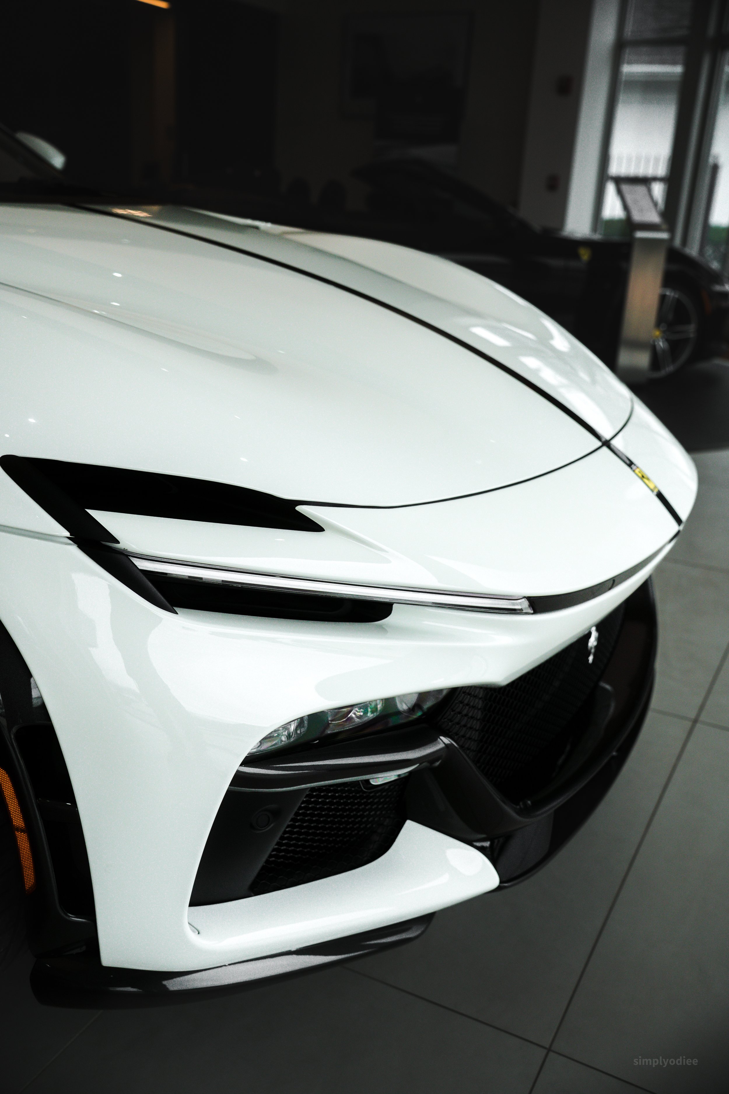 Close-up of the front of a white luxury sports car with sleek headlights and black accents, displayed indoors at a dealership.