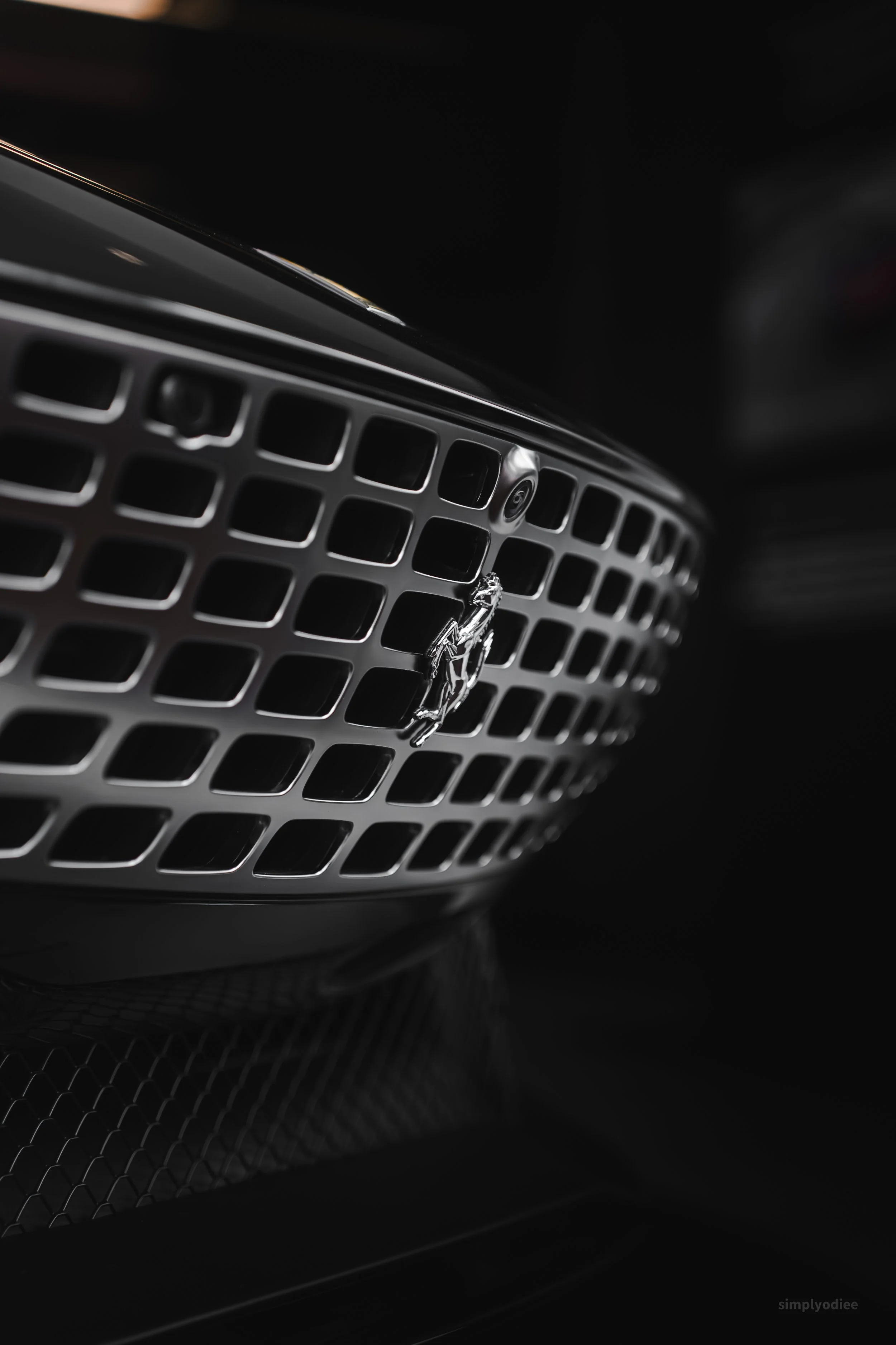 Close-up of a luxury car's front grille with a chrome badge of a horse and a camera above it, in a dark setting.