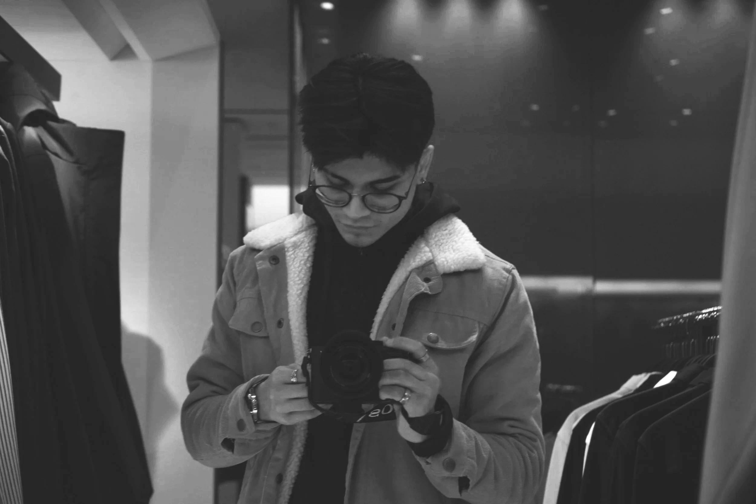Black and white photo of a person with short hair, glasses, wearing a jacket with a shearling collar, taking a selfie in a mirror at a clothing store.