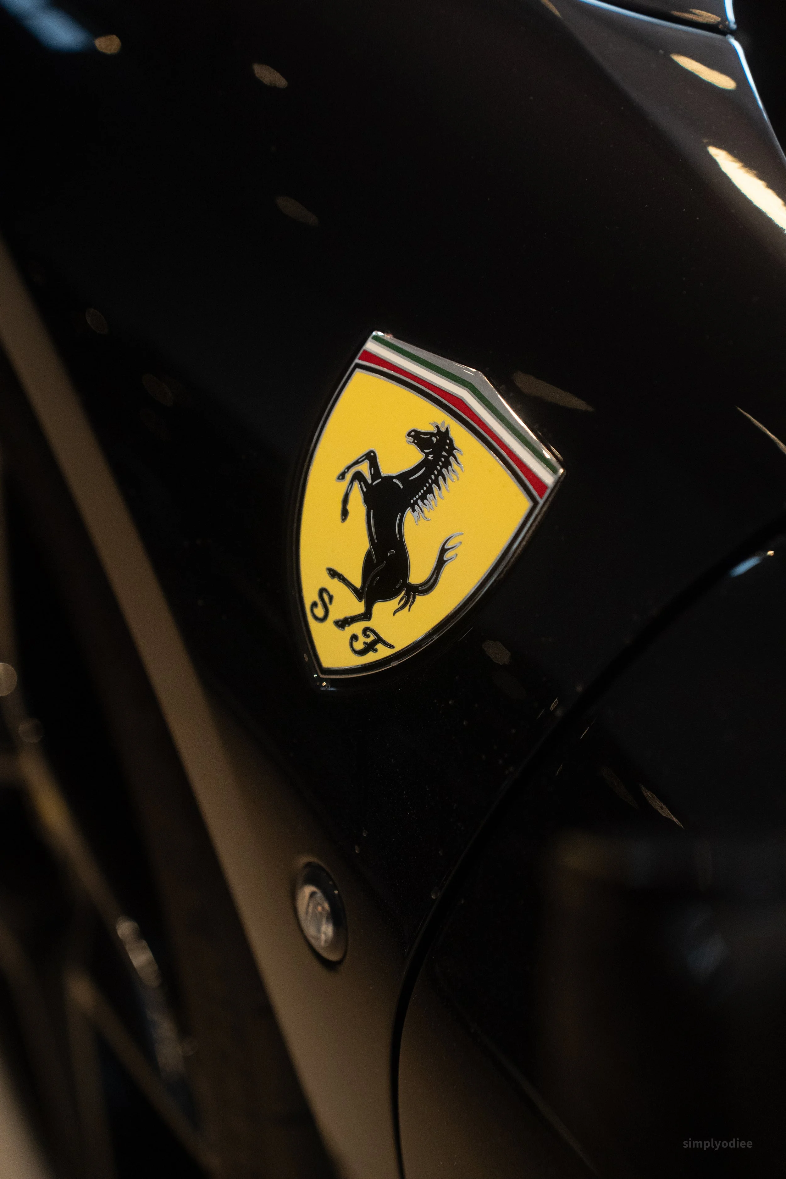 Close-up of a black car's hood featuring a yellow Ferrari badge with a prancing black horse and green, white, and red stripes at the top.