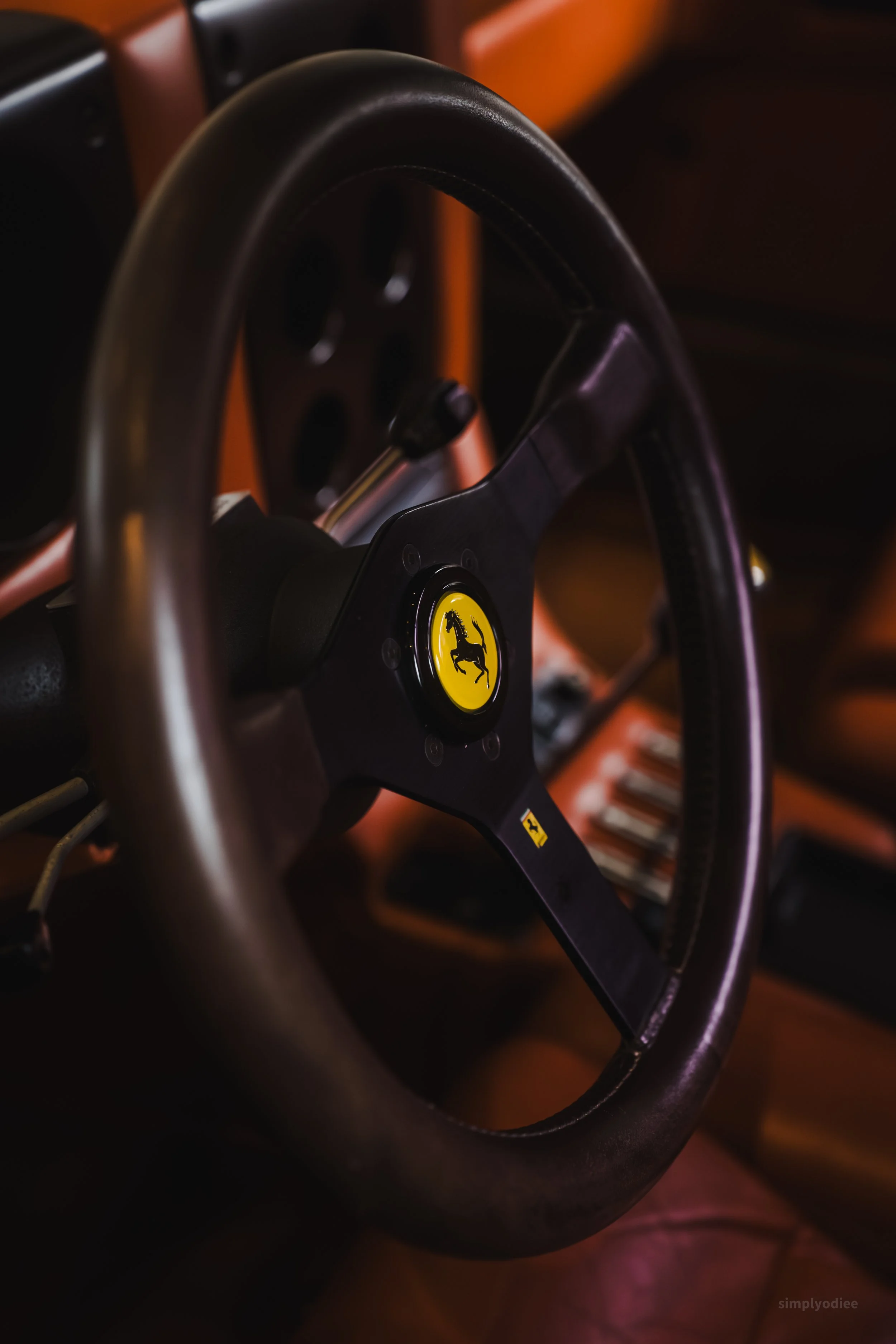 Close-up of a black Ferrari steering wheel with a yellow Ferrari logo in the center, inside a car with brown leather interior.