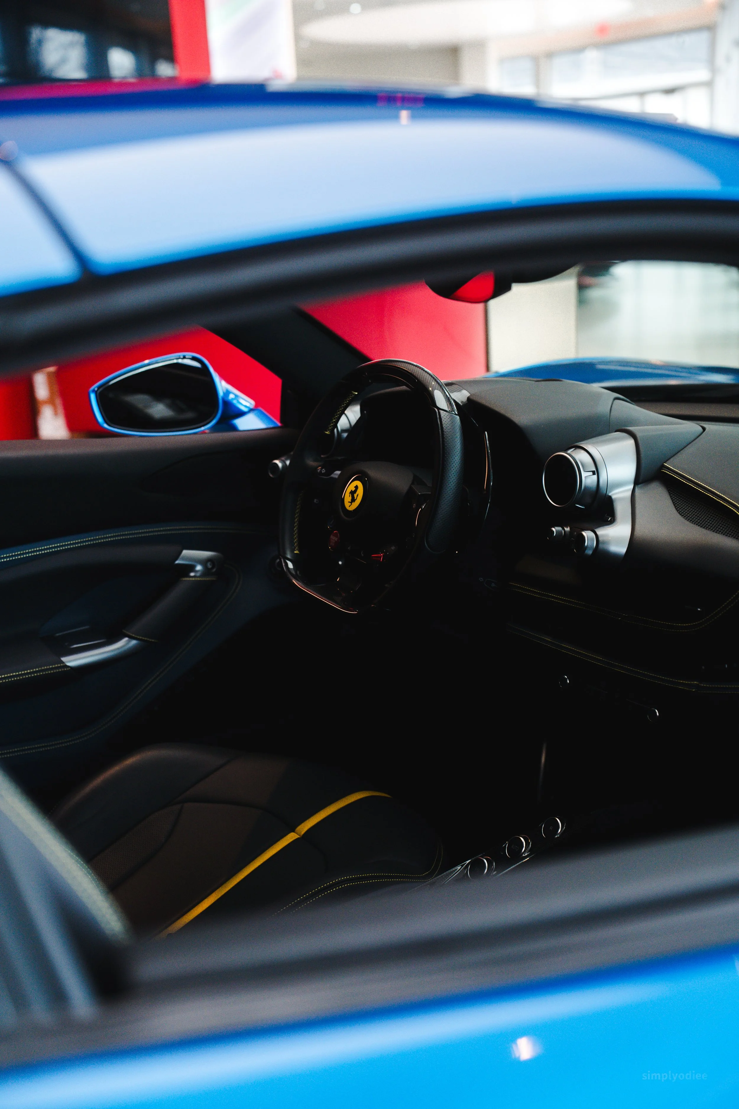 Interior of a blue Ferrari sports car showing steering wheel, dashboard, and seats with yellow stitching, parked indoors.