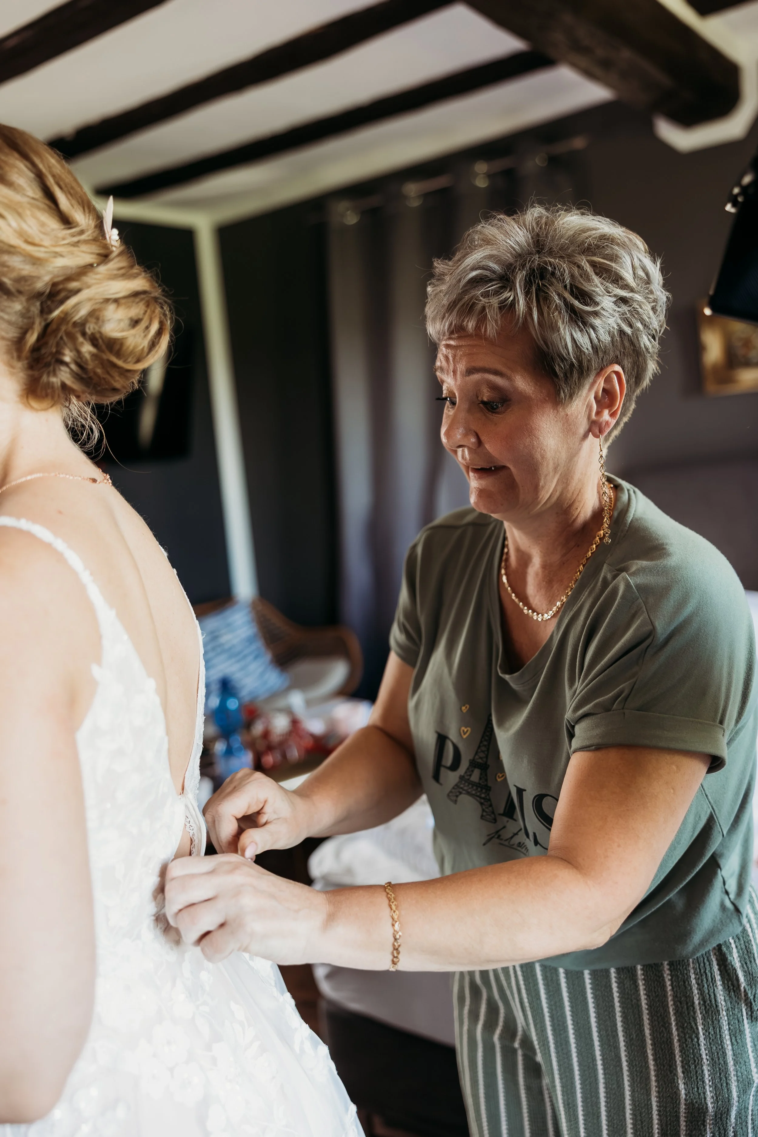 Eine ältere Frau hilft einer jüngeren Frau, die Hochzeitskleid trägt, beim Zubinden des Kleides in einem RaumBraut beim Getting Ready für ihre Hochzeit  auf Mathis Wiesenmühle