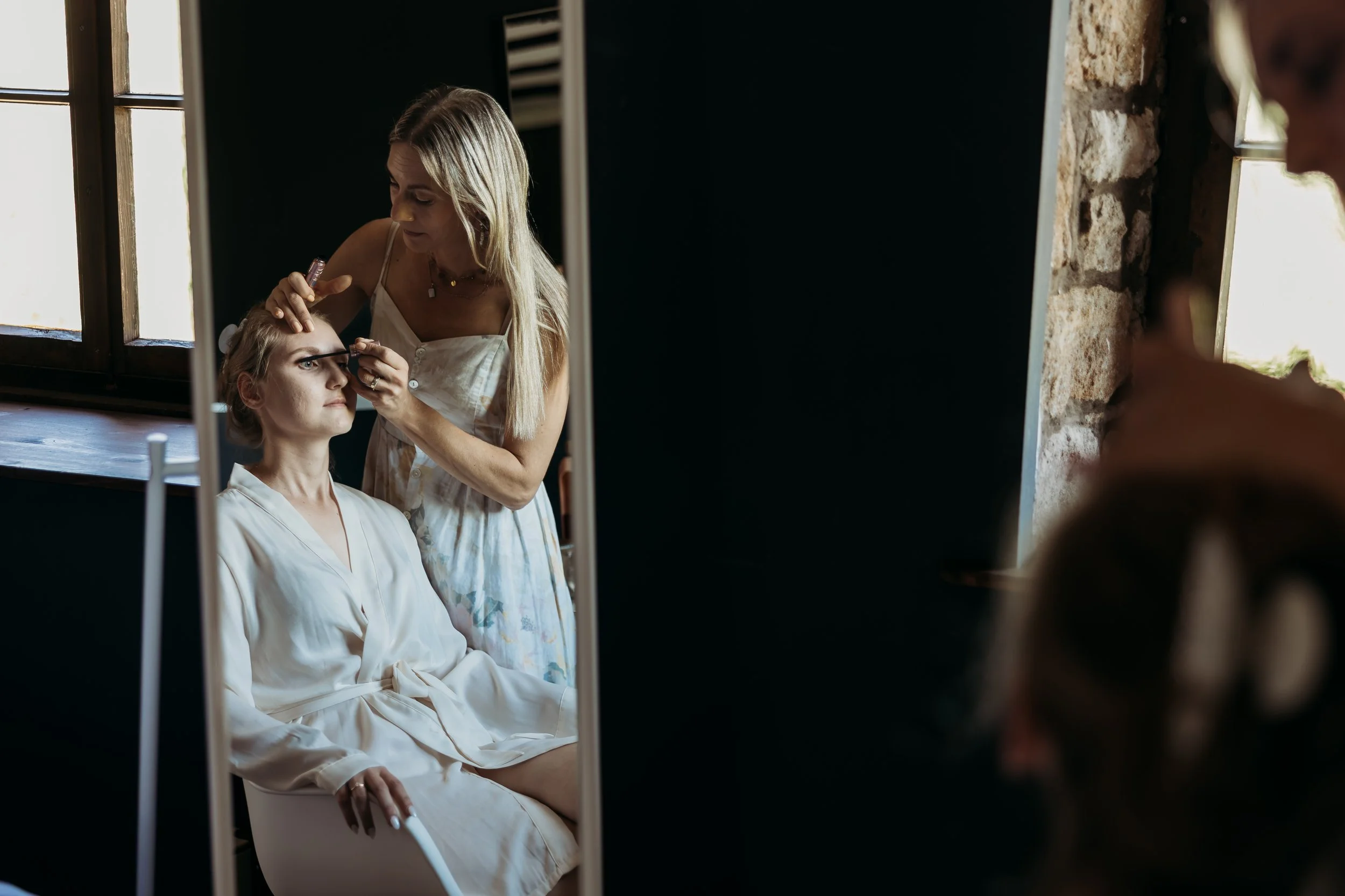 Friseurin schminkt eine Kundin in einem Salon, die auf einem Stuhl sitzt, während sie im Spiegel sieht.