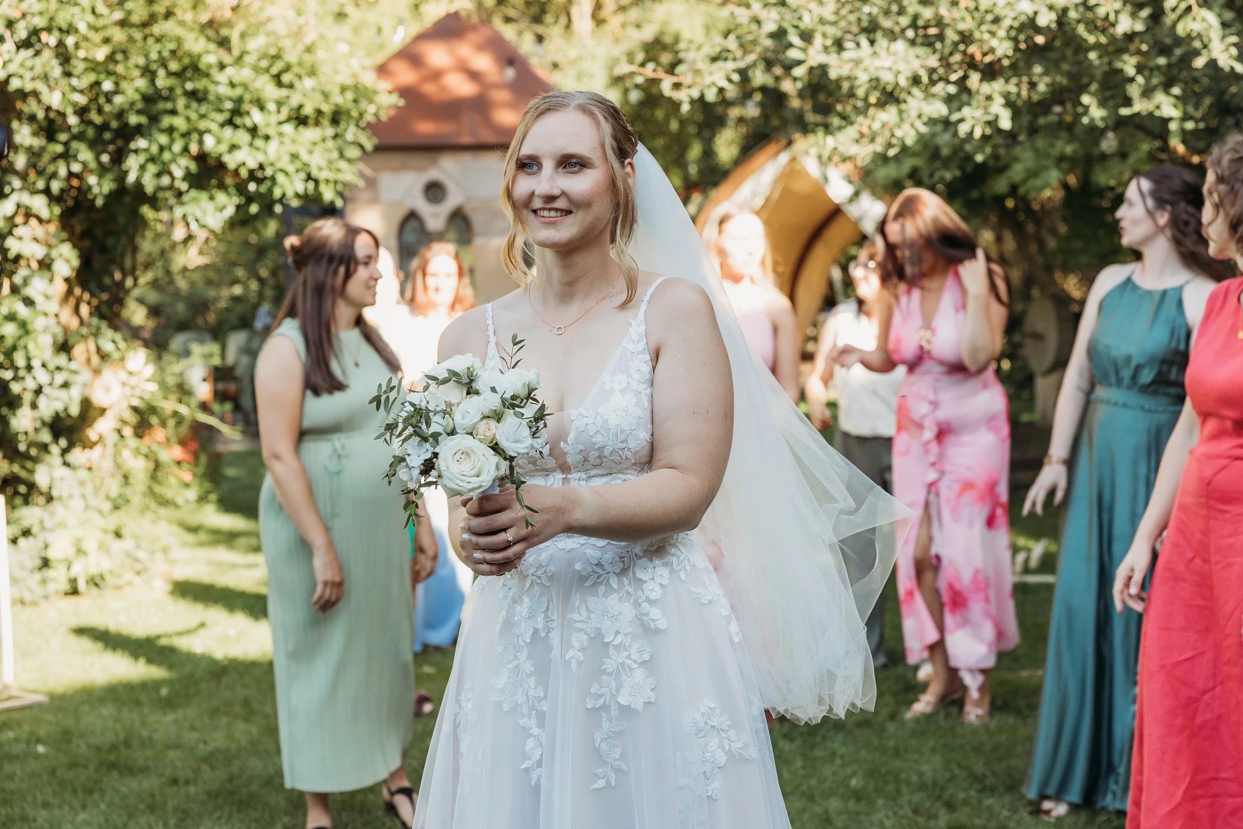 Eine lachende Braut in einem weißen Hochzeitskleid mit Blumenmuster, die einen Blumenstrauß hält, steht im Garten mit mehreren Frauen im Hintergrund.