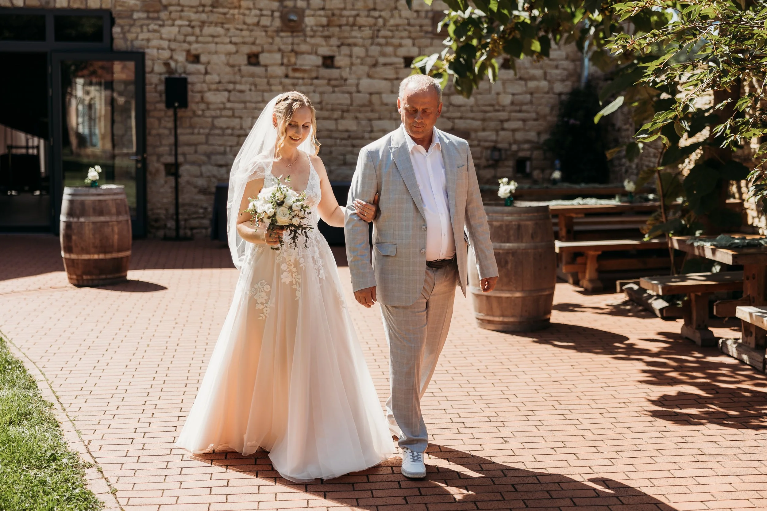 Braut mit Brautstrauß wird von ihrem Vater an ihrem Hochzeitstag im Garten von Mathis Wiesenmühle begleitet.