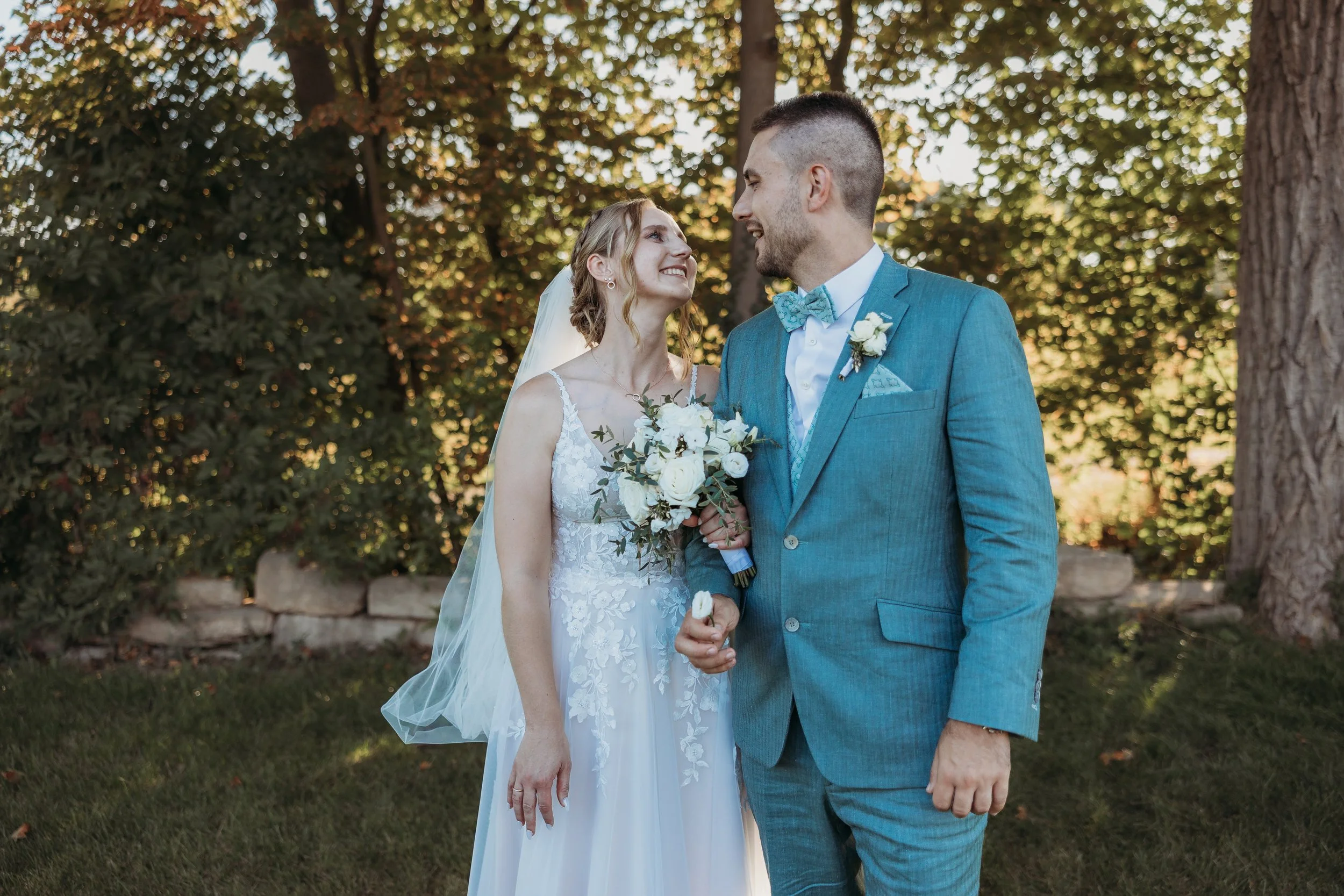 Braut und Bräutigam lächeln sich bei ihrer Hochzeit im Garten an, die Braut trägt ein weißes Kleid und hält einen Blumenstrauß, der Bräutigam trägt einen blauen Anzug mit Fliege.