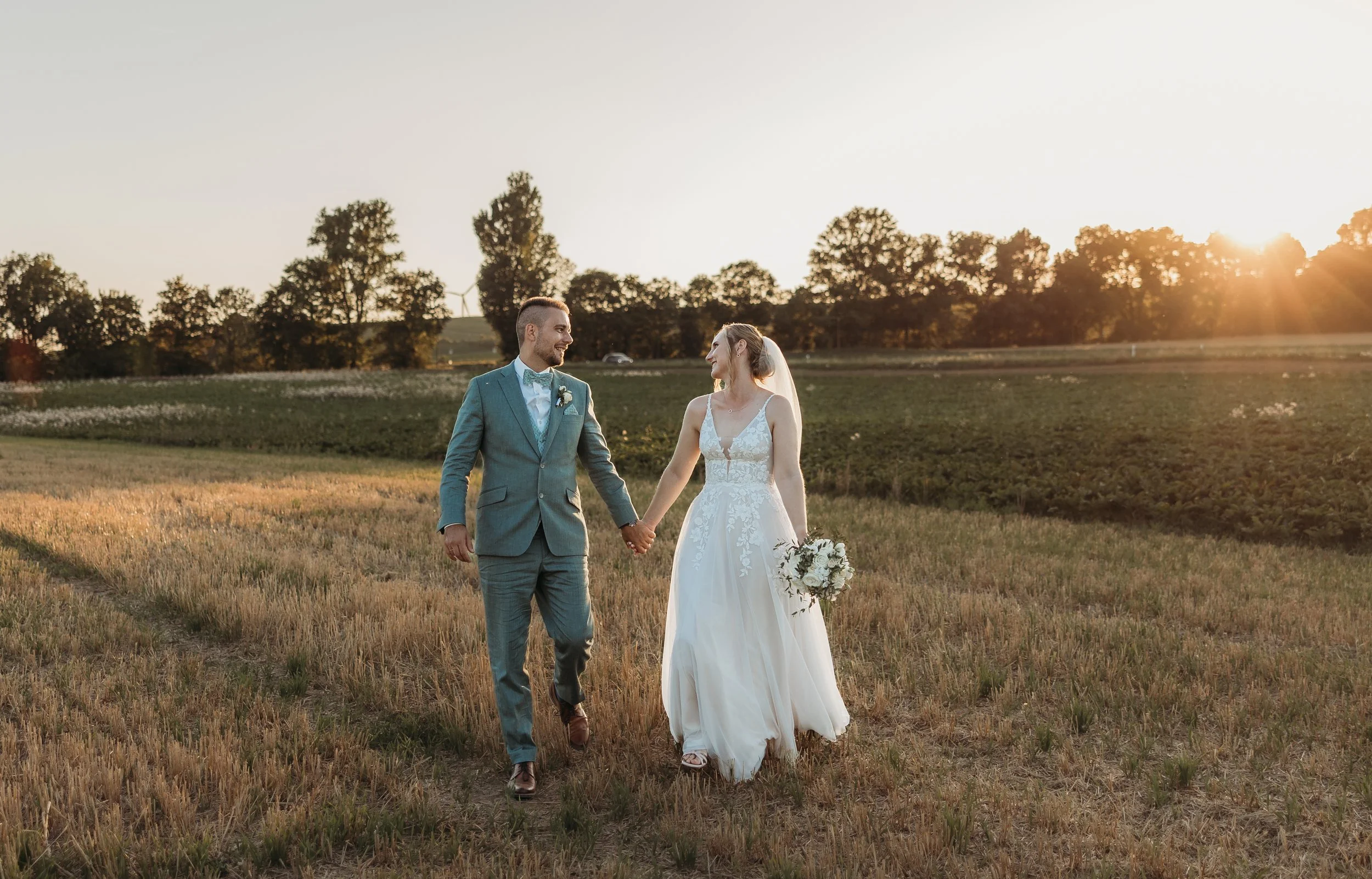 Ein Brautpaar läuft Hand in Hand durch ein Feld bei Sonnenuntergang, die Frau trägt ein weißes Hochzeitskleid und hält einen Blumenstrauß, der Mann trägt einen blauen Anzug.