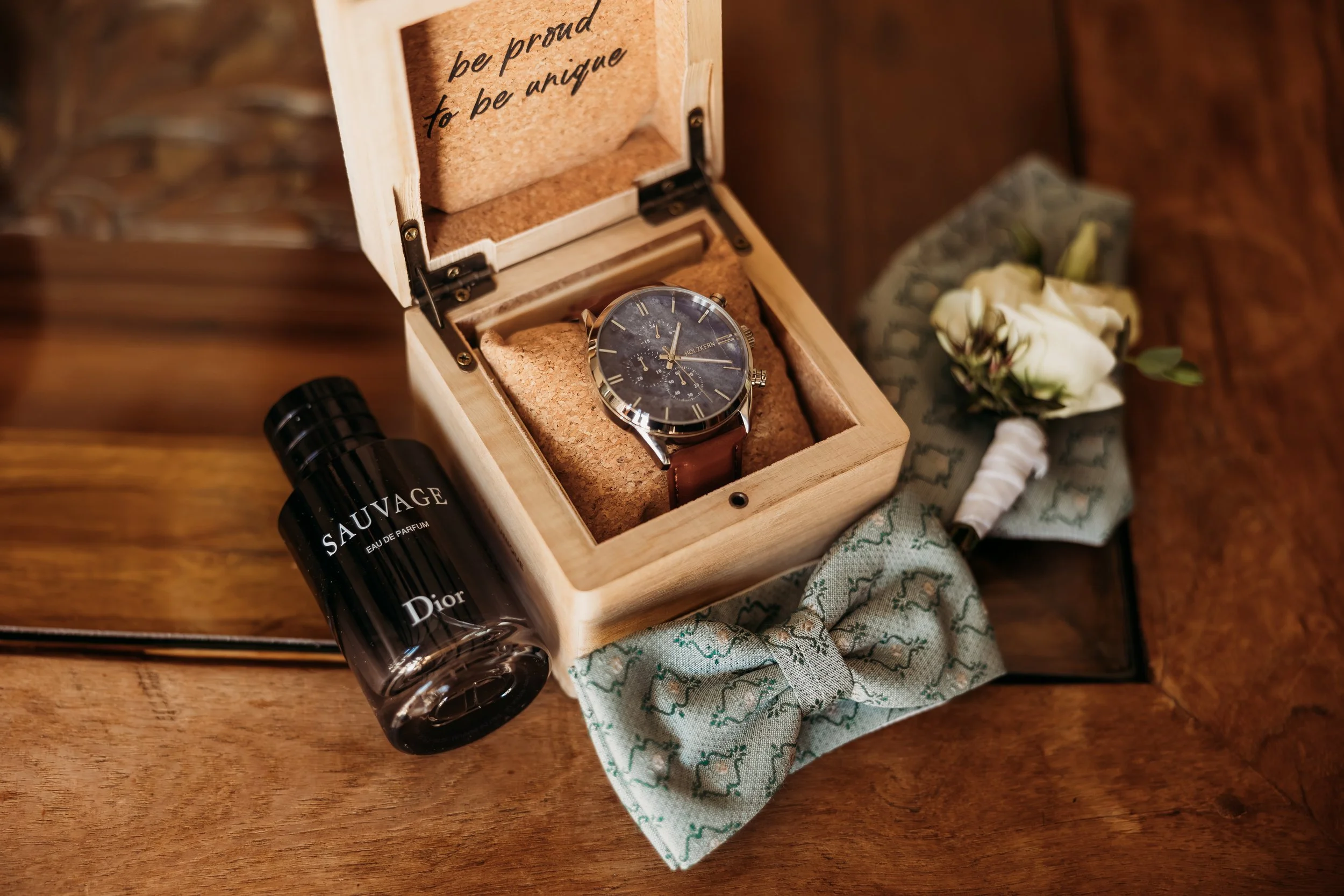 Eine Uhr in einer Holzschatulle, eine weiße Rose mit Schleife, ein schwarzes Parfümfläschchen mit dem Label 'SAUVAGE' von Dior, und ein blauer Stoffband mit Muster liegen auf einem Holztisch. Die Location ist Mathis Wiesenmühle
