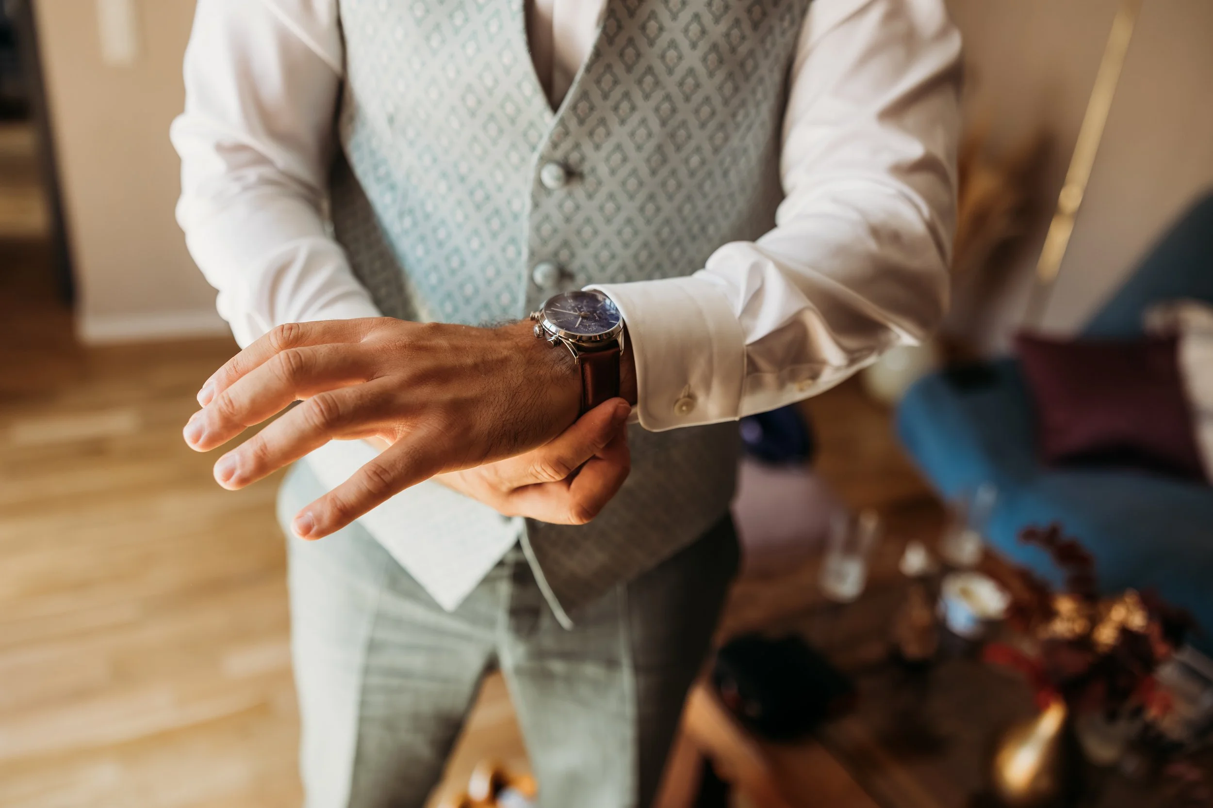 Ein Mann zieht sich eine Armbanduhr an, trägt ein weißes Hemd und eine Weste, im Hintergrund ein Wohnzimmer  auf Mathis Wiesenmühle