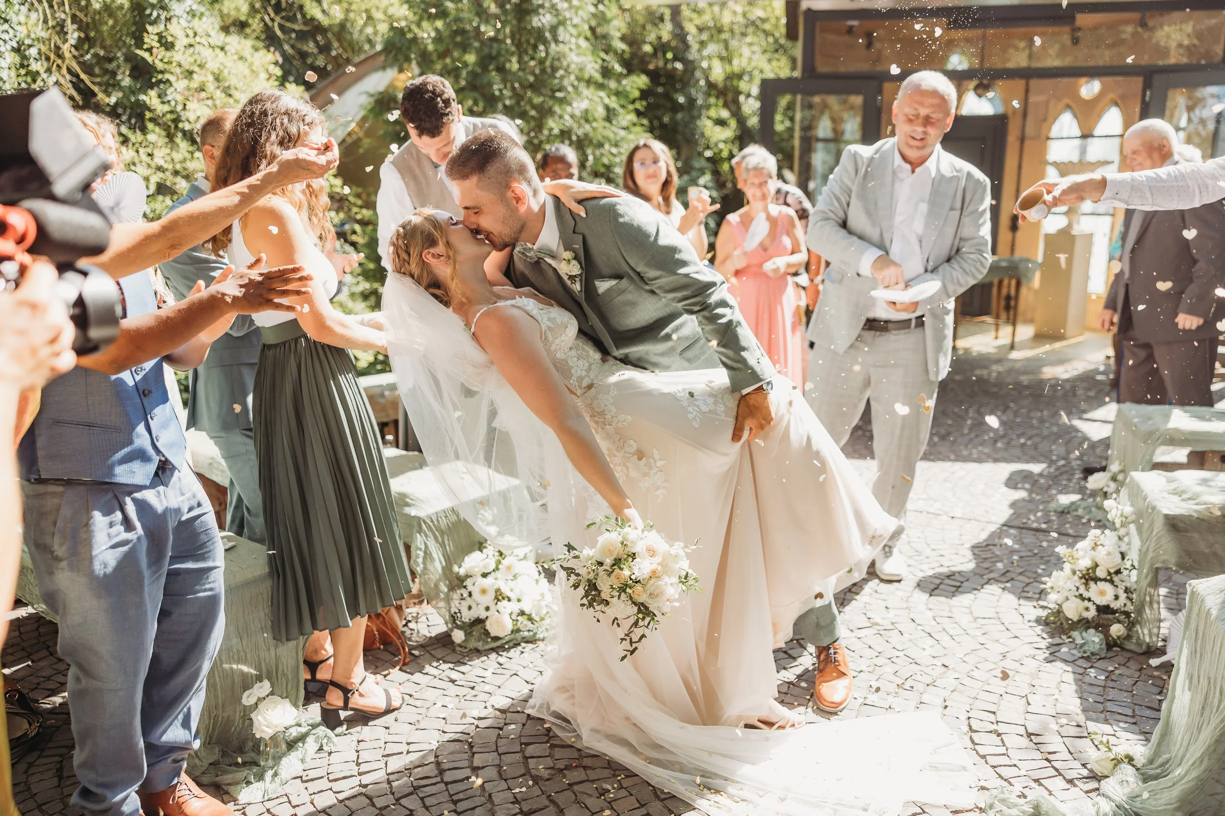 Ein Brautpaar küsst sich bei einer Hochzeit, umgeben von Gästen, die Konfetti werfen, auf einem gepflasterten Platz im Freien, mit bunten Blumen und Dekorationen.