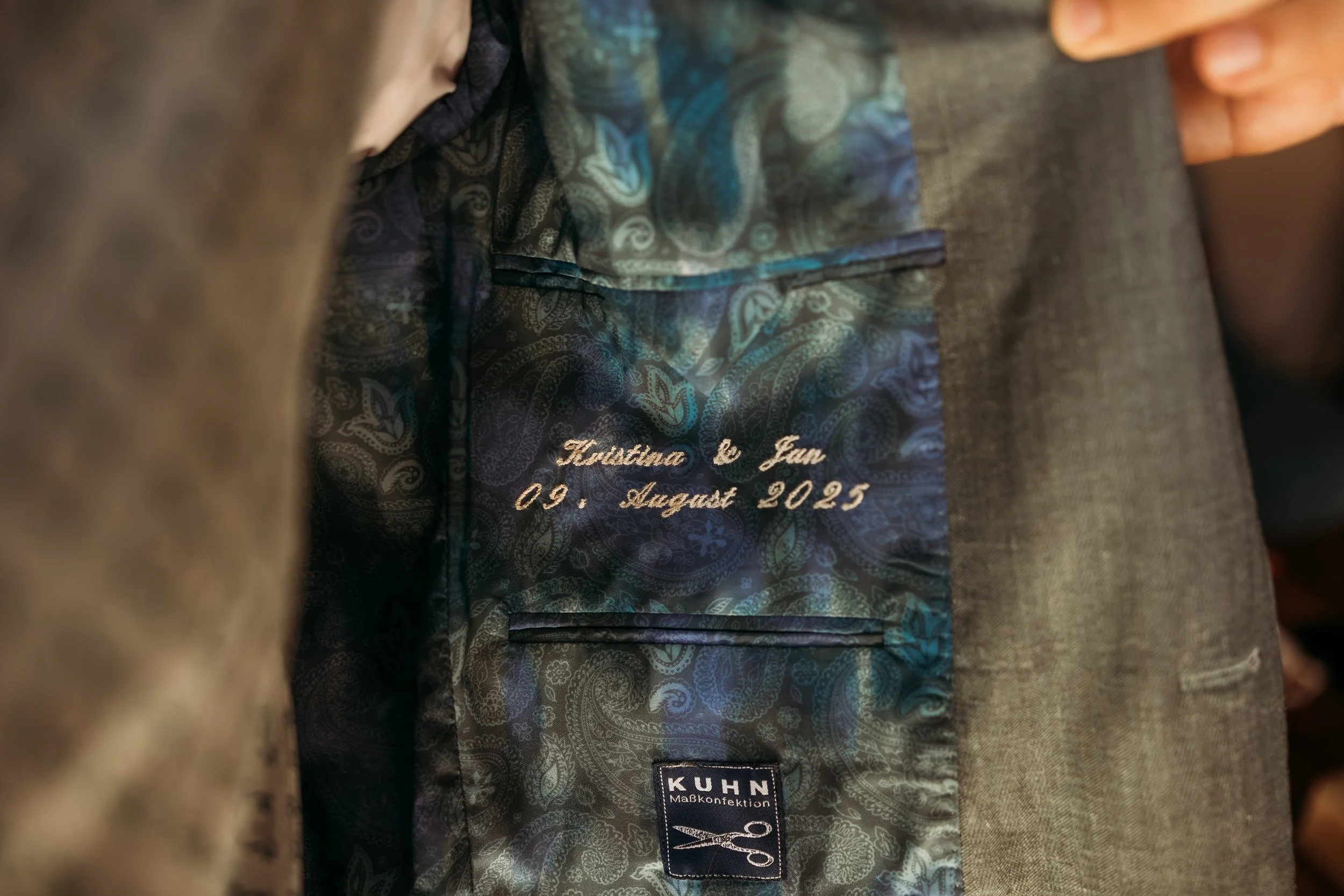 Innenansicht eines Jacketts mit Stickerei, die den Namen 'Kristina & Jan' und das Datum '09. August 2025' zeigt, sowie einem Etikett mit der Marke 'KUHN Maßkonfektion'. Die Hochzeit findet  auf Mathis Wiesenmühle statt
