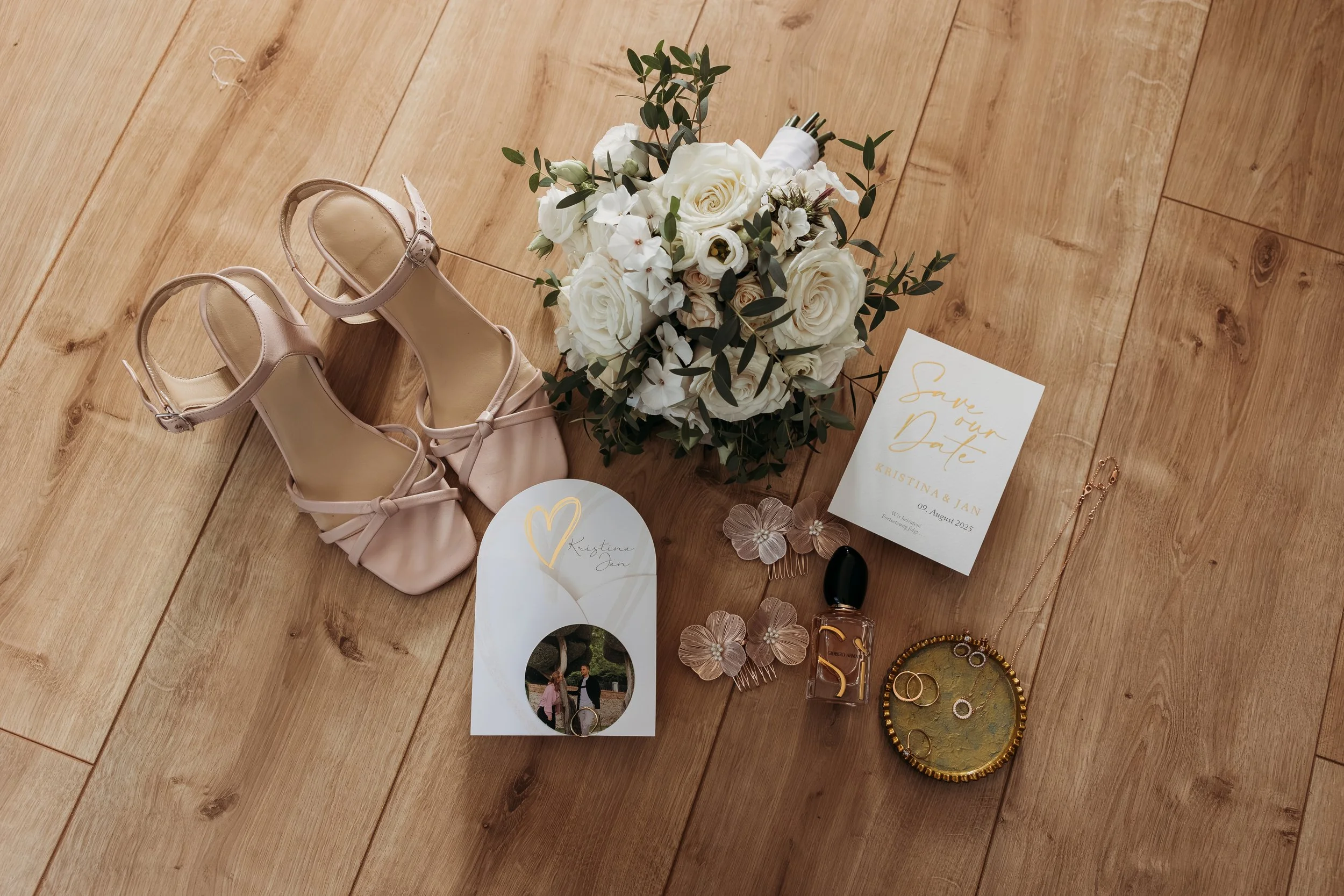Brautaccessoires und Hochzeitsdekoration auf Holzboden, darunter ein Brautschuh, Blumenstrauß, Hochzeitskarte, Foto, Schmuck und Parfümauf Mathis Wiesenmühle