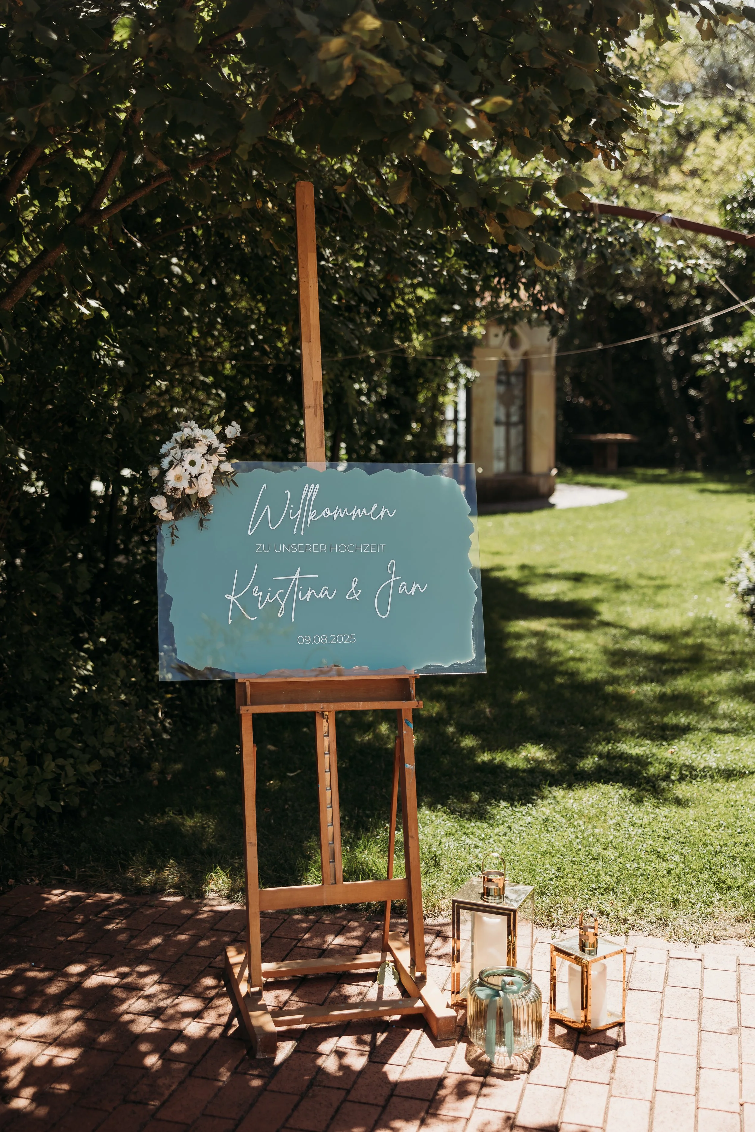 Ein Willkommensschild auf einer easel bei einer Hochzeit mit weißen Blumen und einem Garten im Hintergrund.