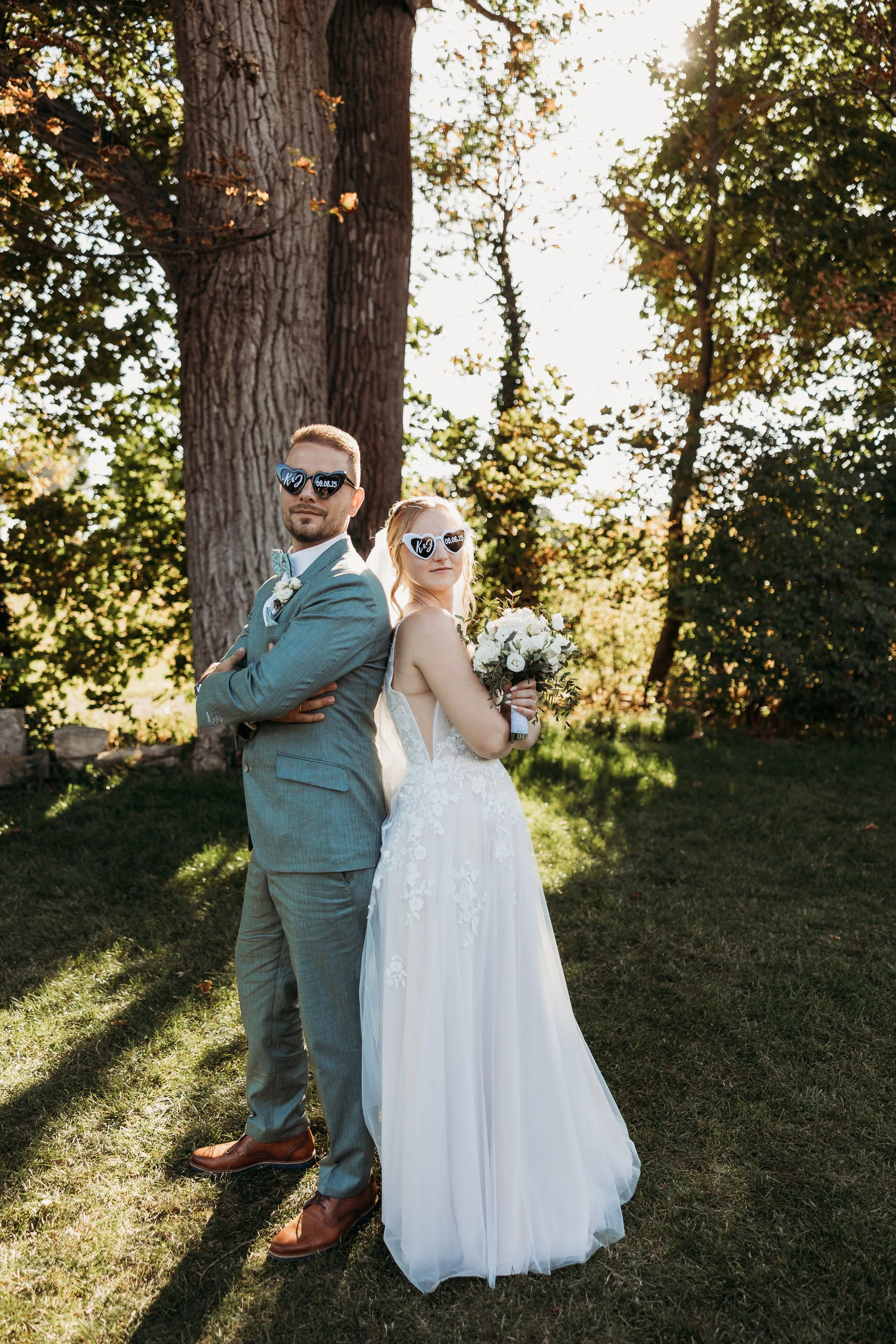 Ein Brautpaar bei ihrer Hochzeit, stehend Rücken an Rücken unter einem Baum, beide tragen Sonnenbrillen, das Brautpaar hält einen Blumenstrauß, die Szene ist im Freien bei Sonnenlicht.