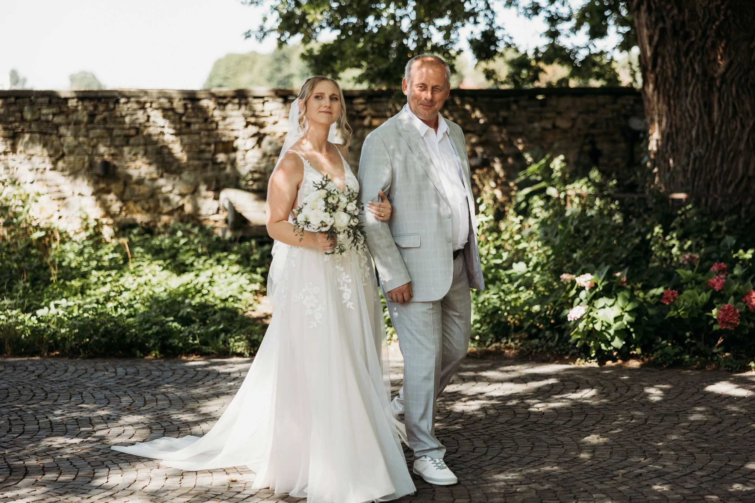 Braut in weißem Brautkleid mit Blumenstrauß, neben älterem Mann im hellgrauen Anzug bei Hochzeit im Freien auf Mathis Wiesenmühle