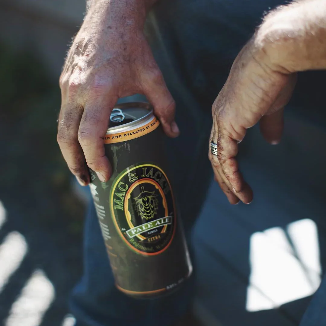 Person holding a can of Mac & Jack's Pale Ale beer with both hands, outdoors in sunlight.