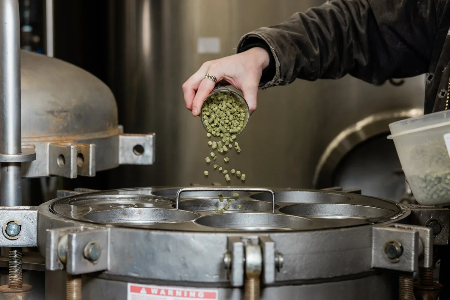 A person pouring green hops into a fermenter.