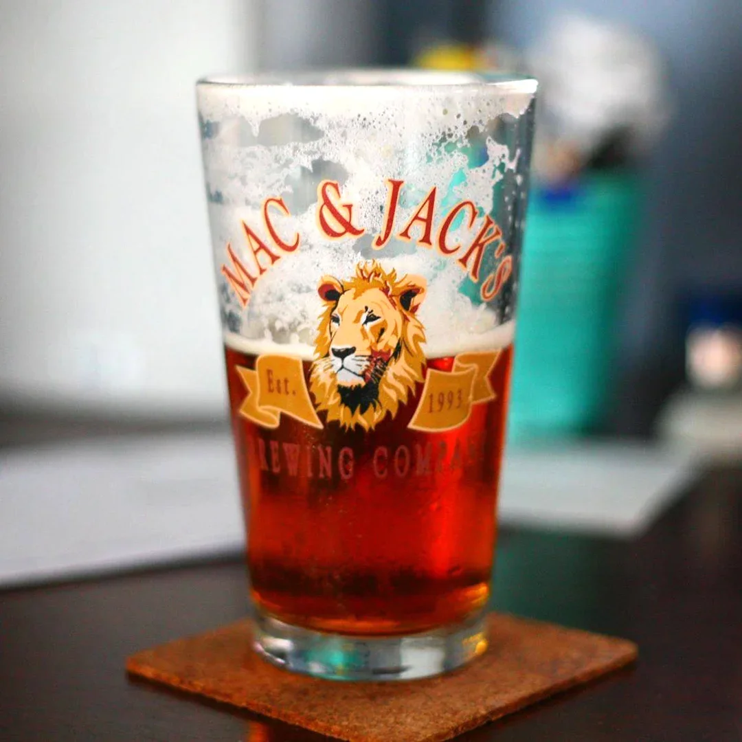 A glass of beer with the logo of Mac & Jack's Brewing Company featuring a lion's head, on a dark surface with a blurred background.