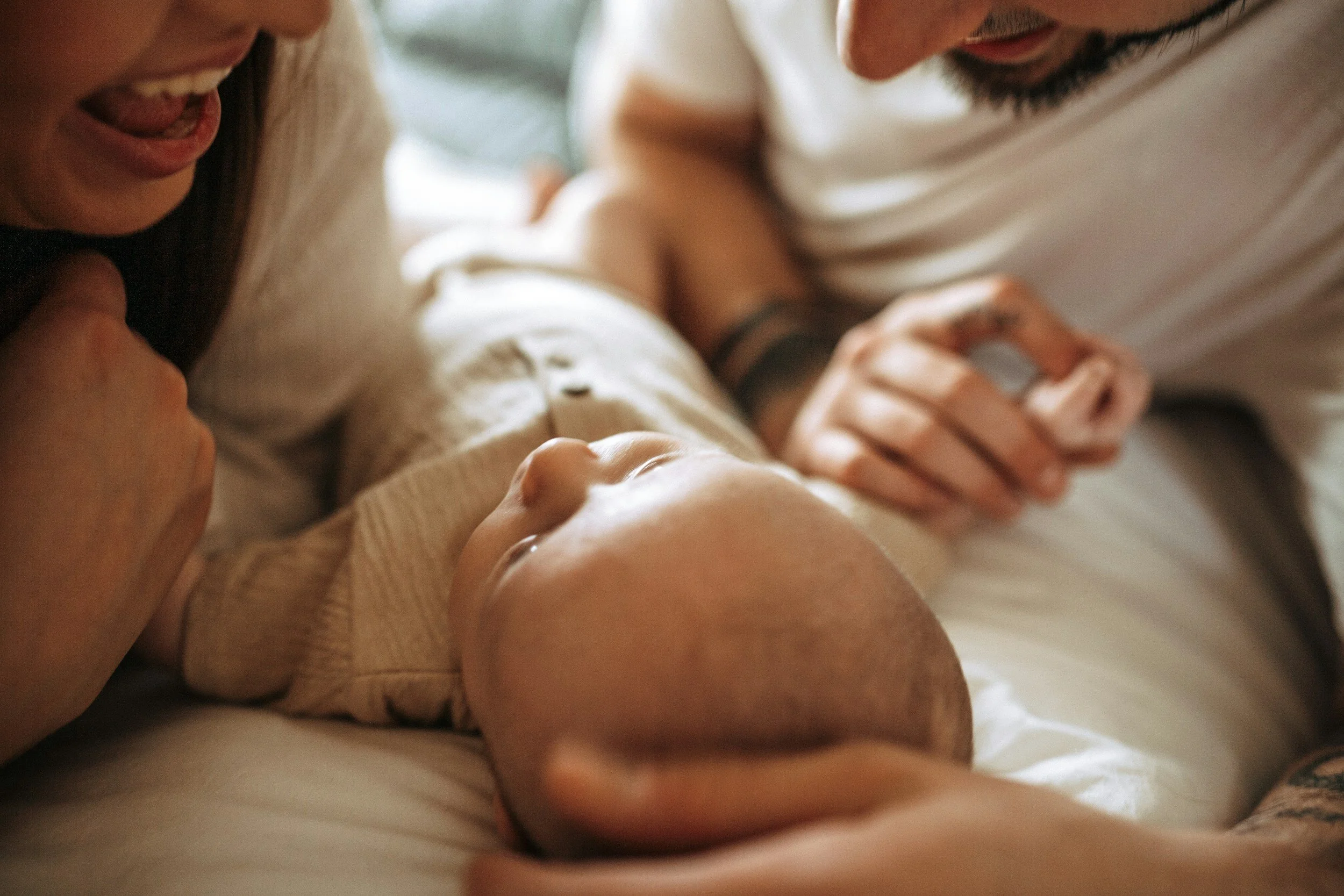 A close-up of a family scene showing an adult couple and a baby, with the baby lying on a bed. The woman, on the left, is smiling and leaning towards the baby. The man, on the right, is looking at the baby, with hands clasped together. The baby is lying on its back, looking up, with a peaceful expression.