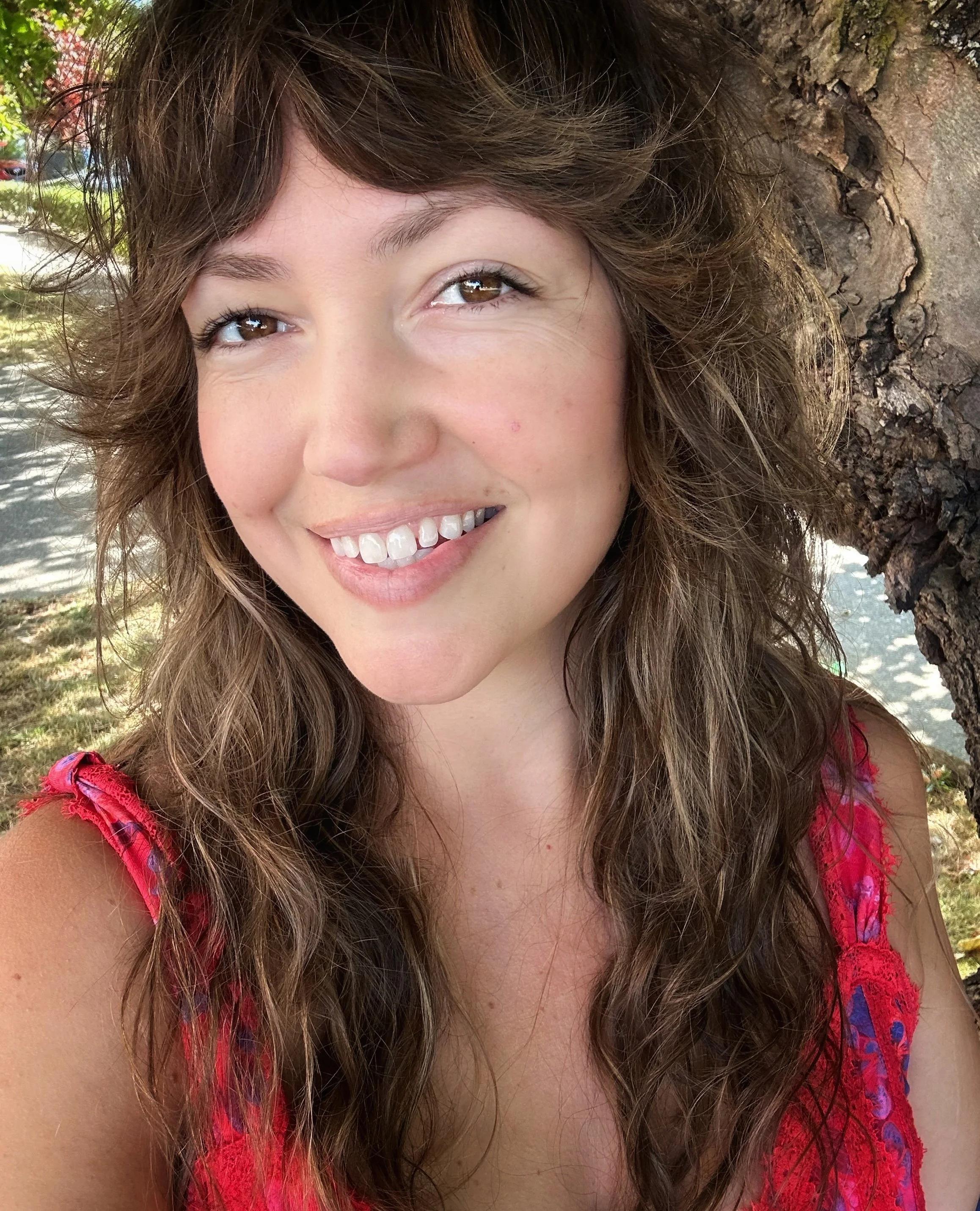A smiling woman with long, wavy brown hair leaning against a tree outdoors, wearing a pink and red sleeveless top.