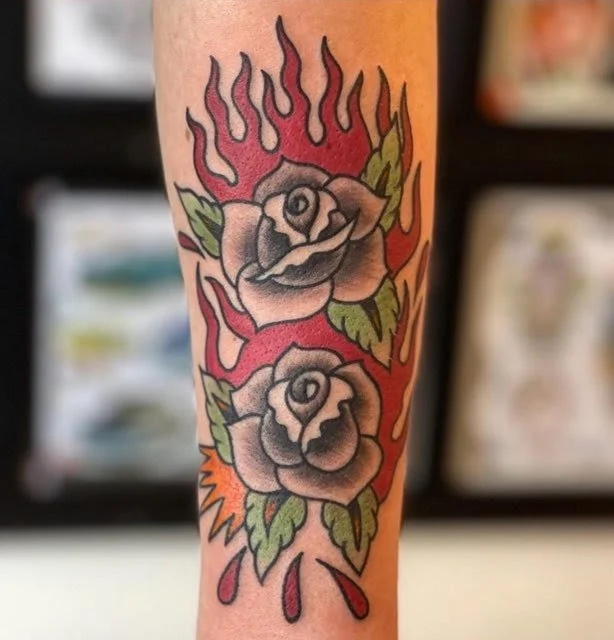 Tattoo of two roses with green leaves surrounded by red and orange flames on a person's arm.