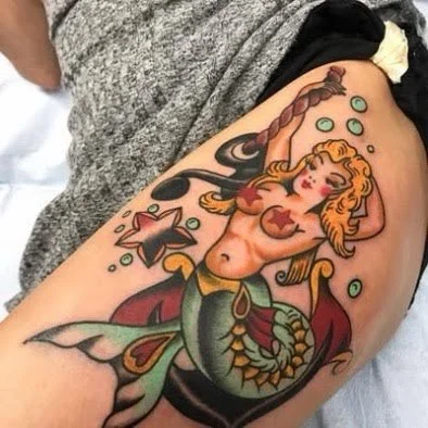 Colorful tattoo of a mermaid with red hair, starfish, seashells, and bubbles on a person's upper arm.