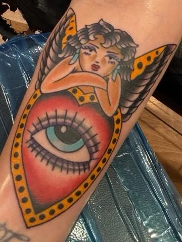 Tattoo of a woman with butterfly wings resting her head on her arms, emerging from a large, stylized eye with eyelashes, surrounded by a heart-shaped border with yellow and black spots.